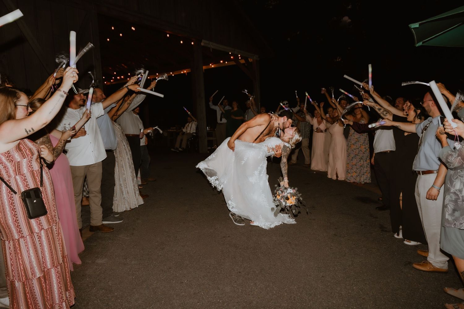 Wedding couple shares a dip kiss during sparkler send-off exit surrounded by cheering guests at night.