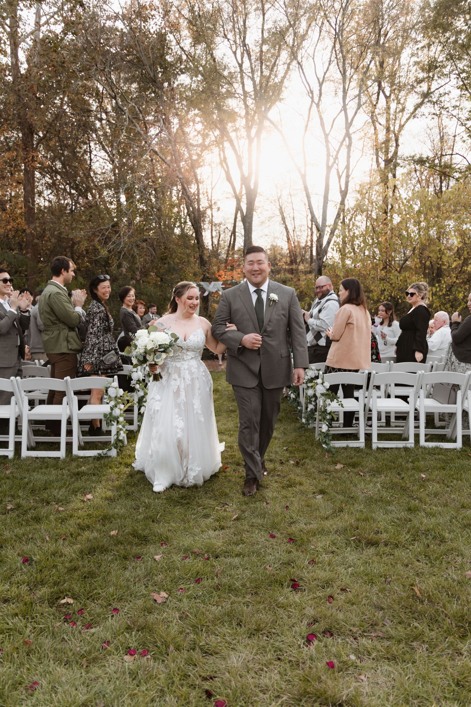 Bride and groom walk down outdoor wedding aisle surrounded by guests and autumn trees at golden hour.