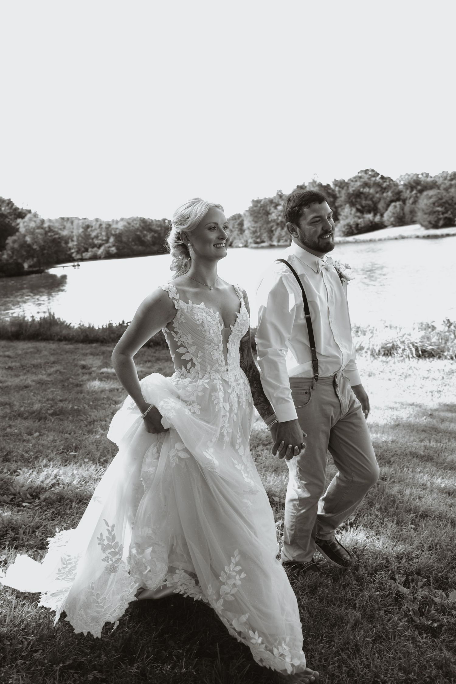 Black and white photo of a smiling bride in lace gown and groom in suspenders walking hand in hand by a lake.