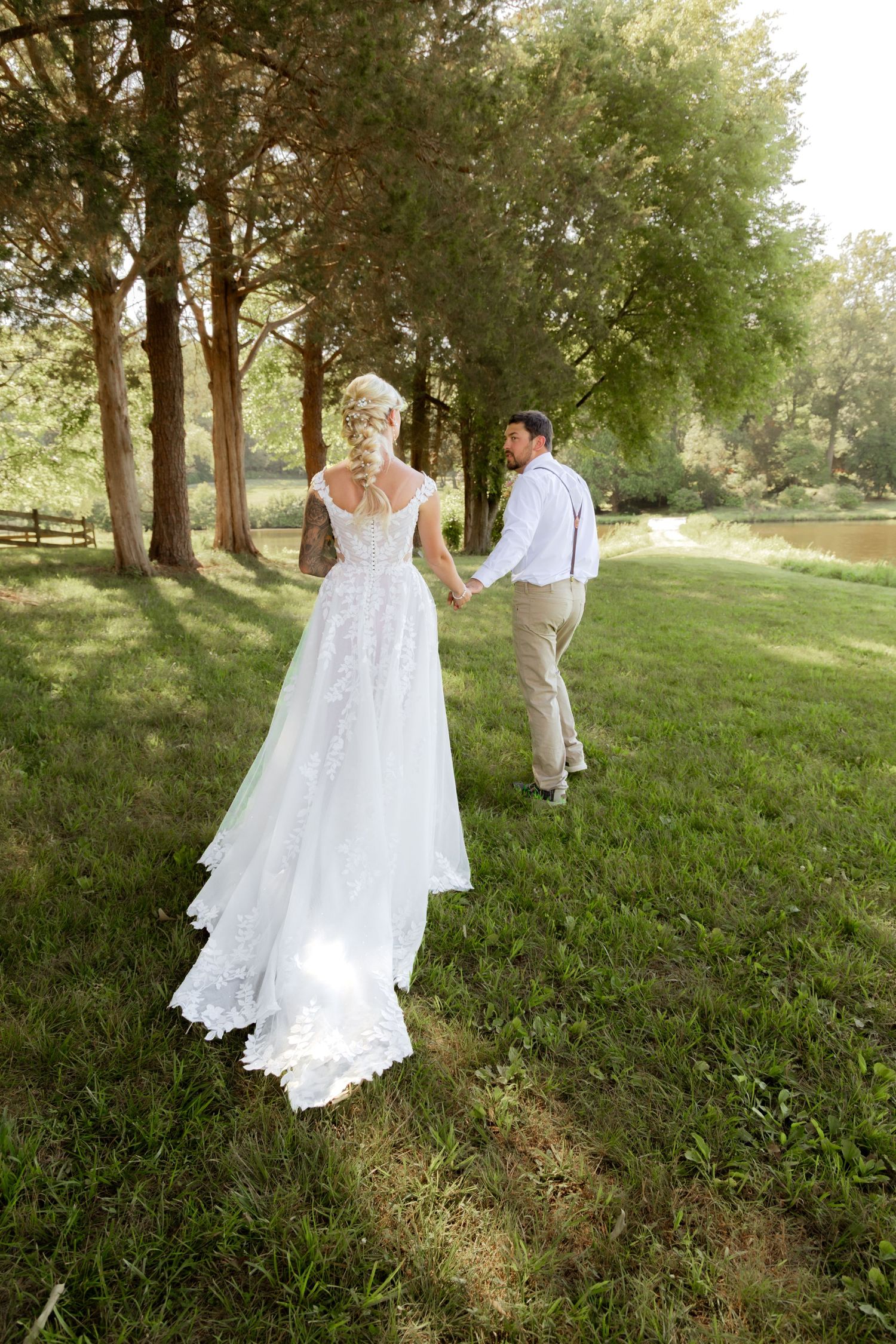Bride and groom holding hands walking through lush green park, bride wearing elegant lace wedding gown with long train.