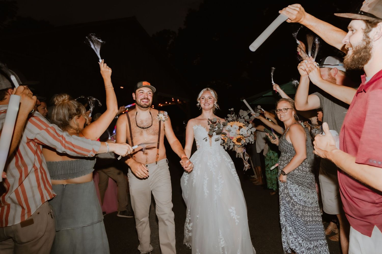 Bride and groom walk through a glowstick tunnel surrounded by cheering wedding guests at a nighttime outdoor reception.