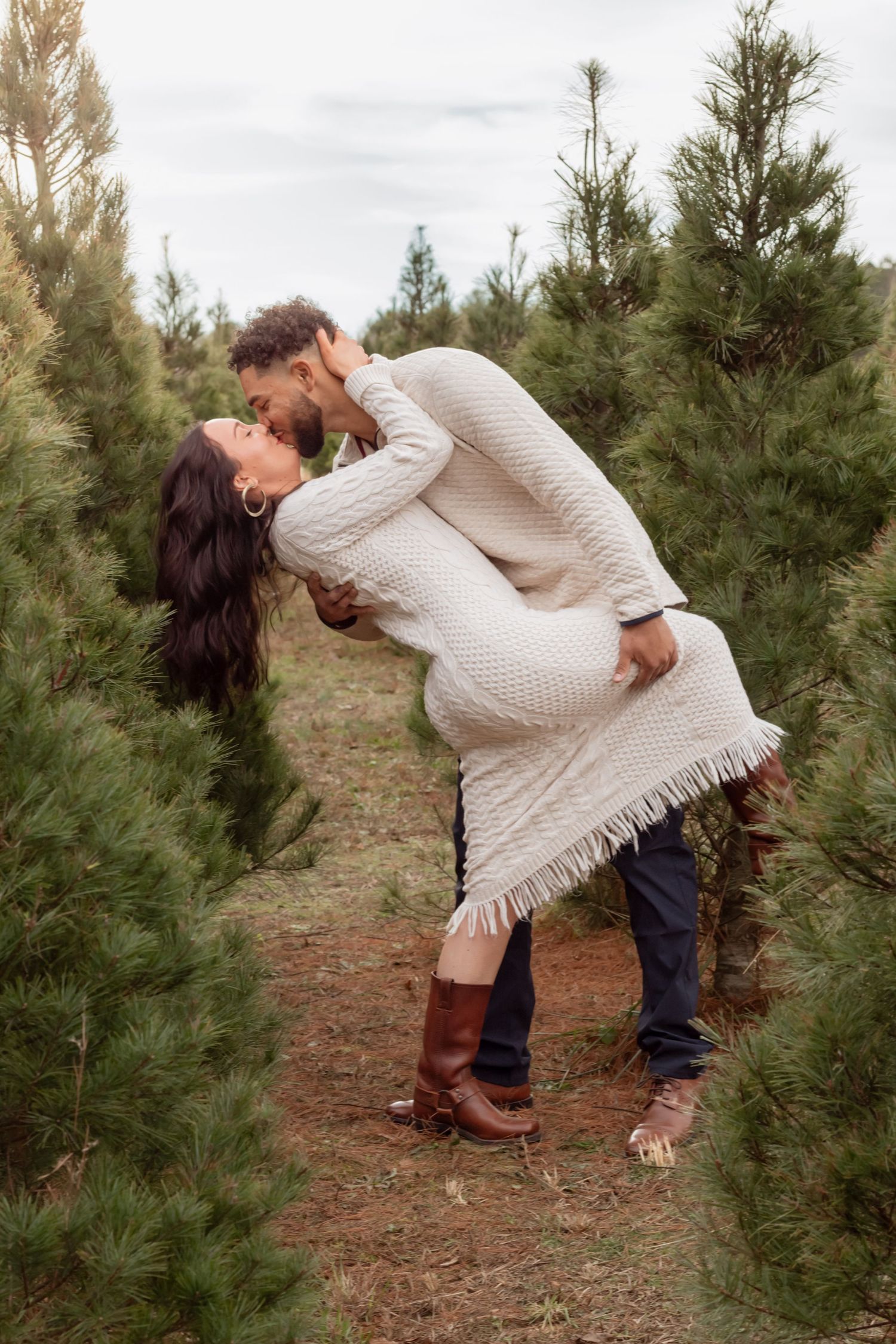 Person in knit sweater bending down among evergreen trees in a forest setting.