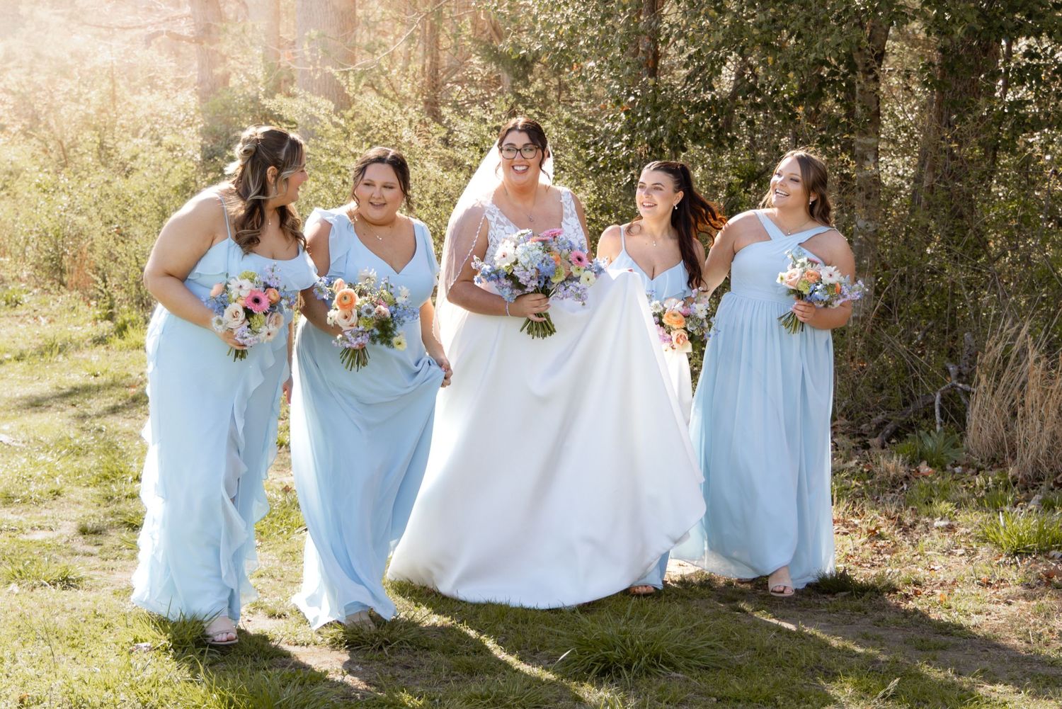 Bride in white gown walks with four bridesmaids in light blue dresses, holding colorful bouquets outdoors.