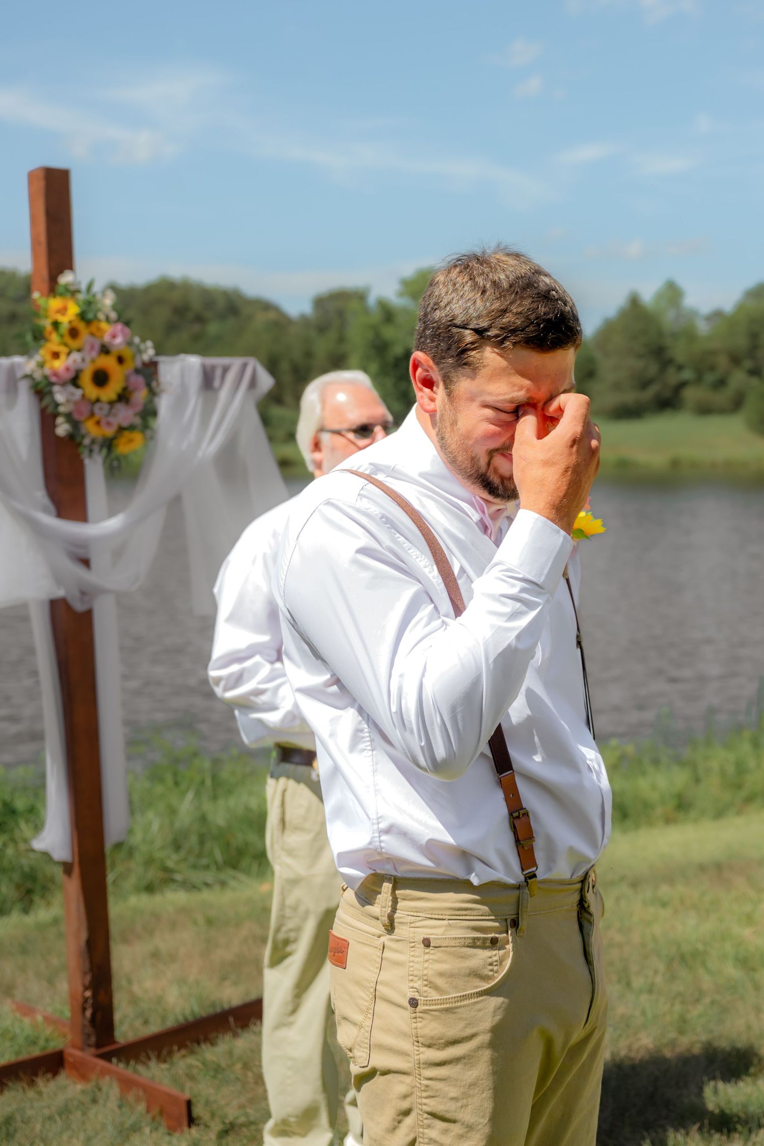 Emotional groom wiping tears at outdoor wedding ceremony by lake, wearing white shirt and suspenders near floral cross.