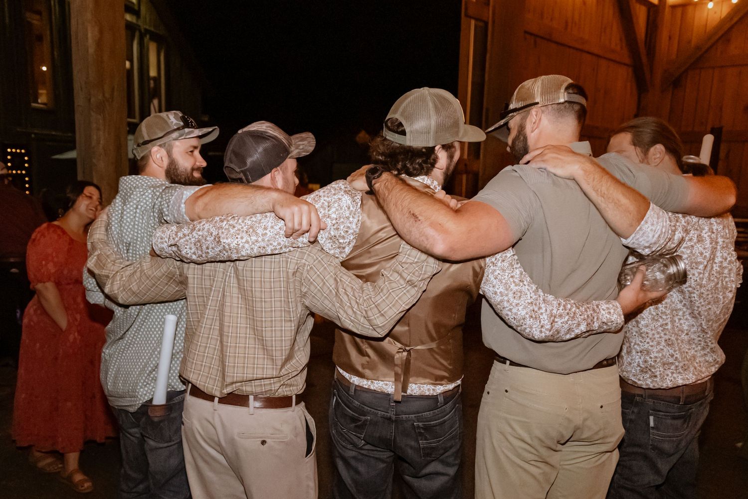 Group of groomsmen in matching outfits hugging together at an outdoor wedding reception at night.