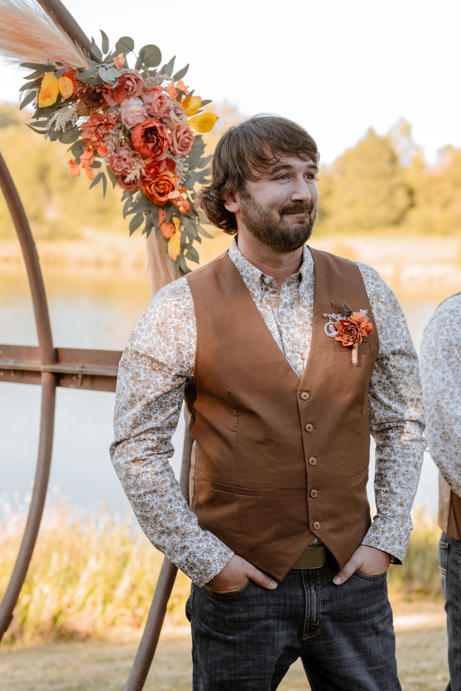 Groom in brown vest and patterned shirt poses against autumn lakeside backdrop.