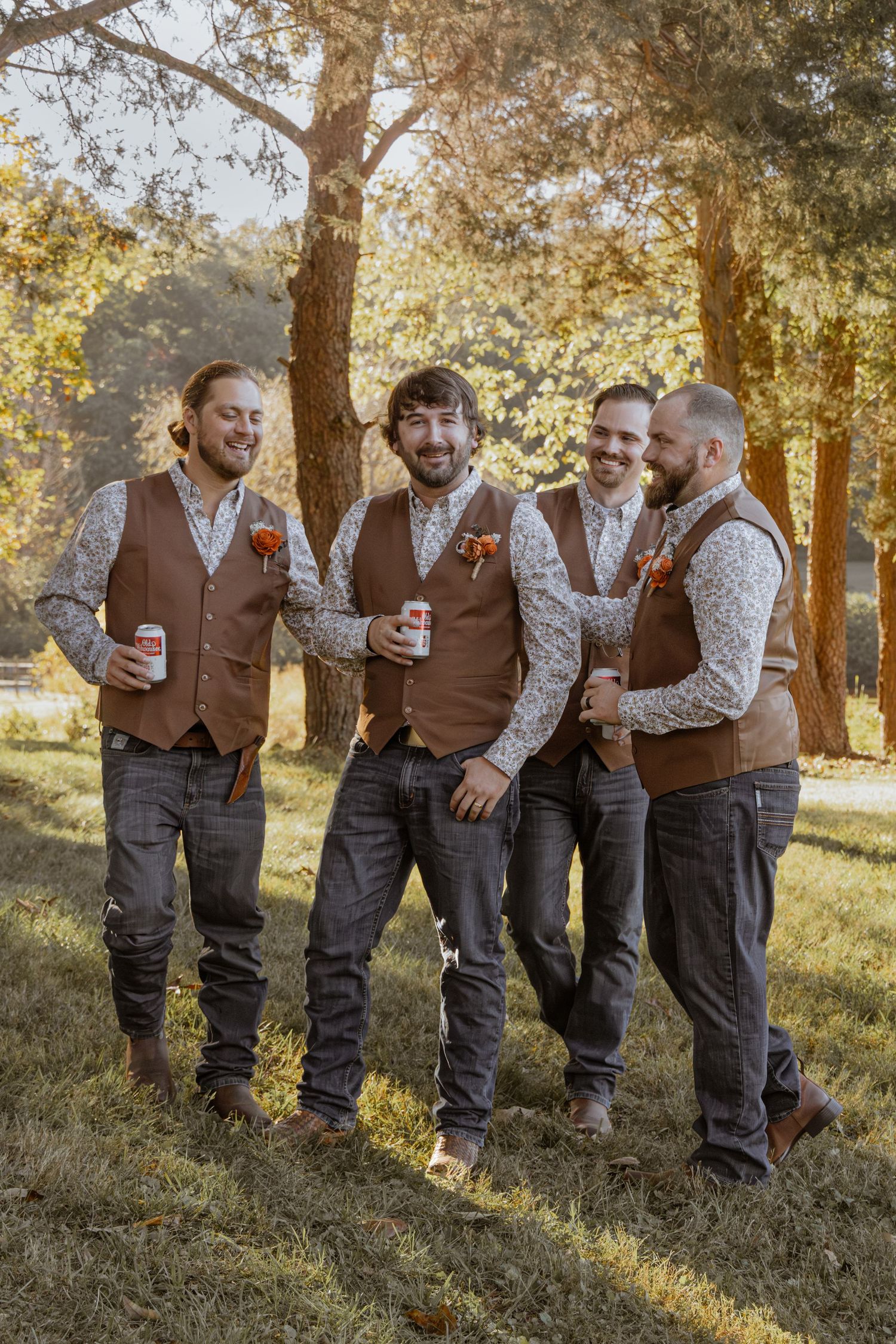 Groomsmen in matching vests and patterned shirts share laughs under trees at sunset.