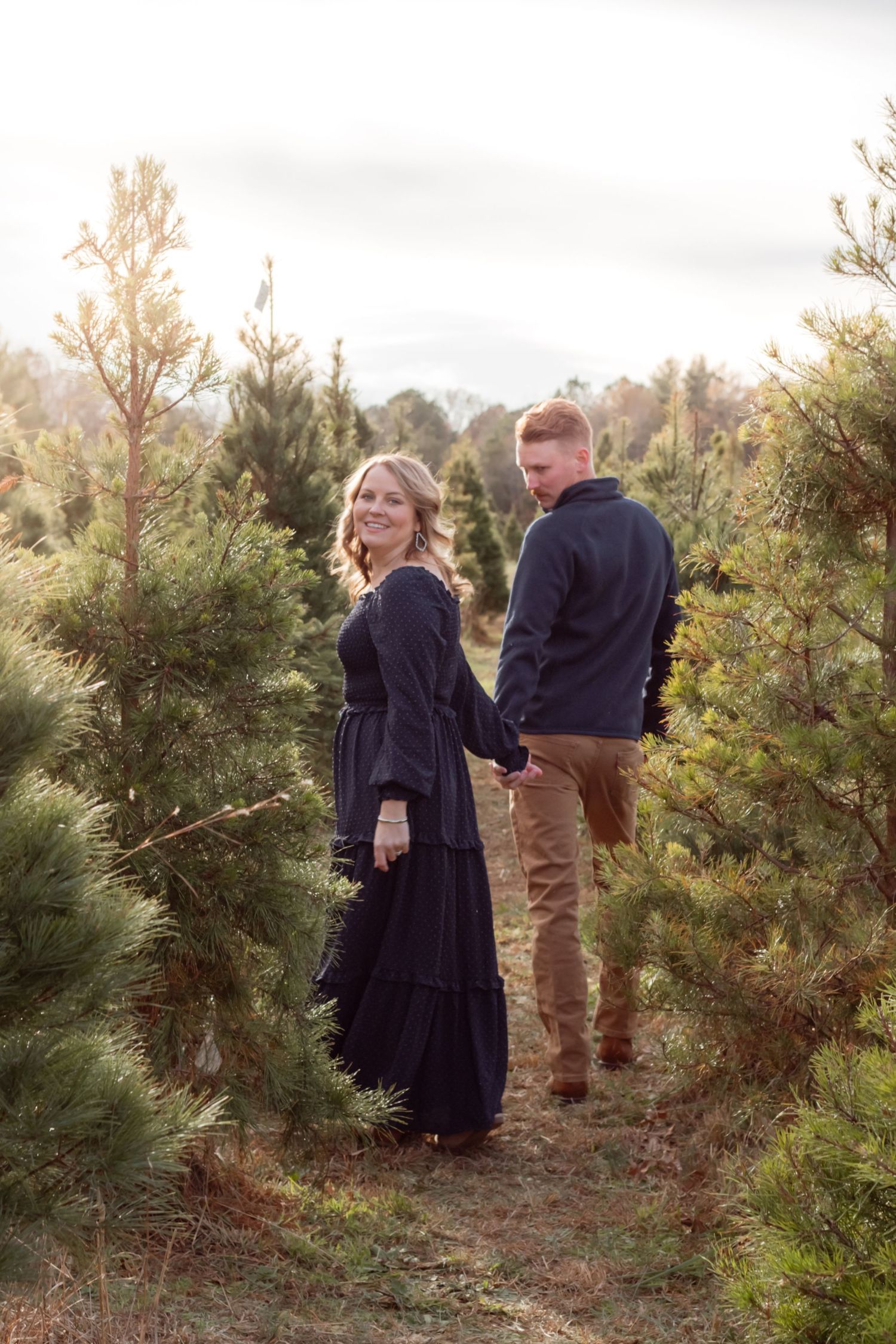 Couple in navy and brown outfits pose among pine trees during a photo session.