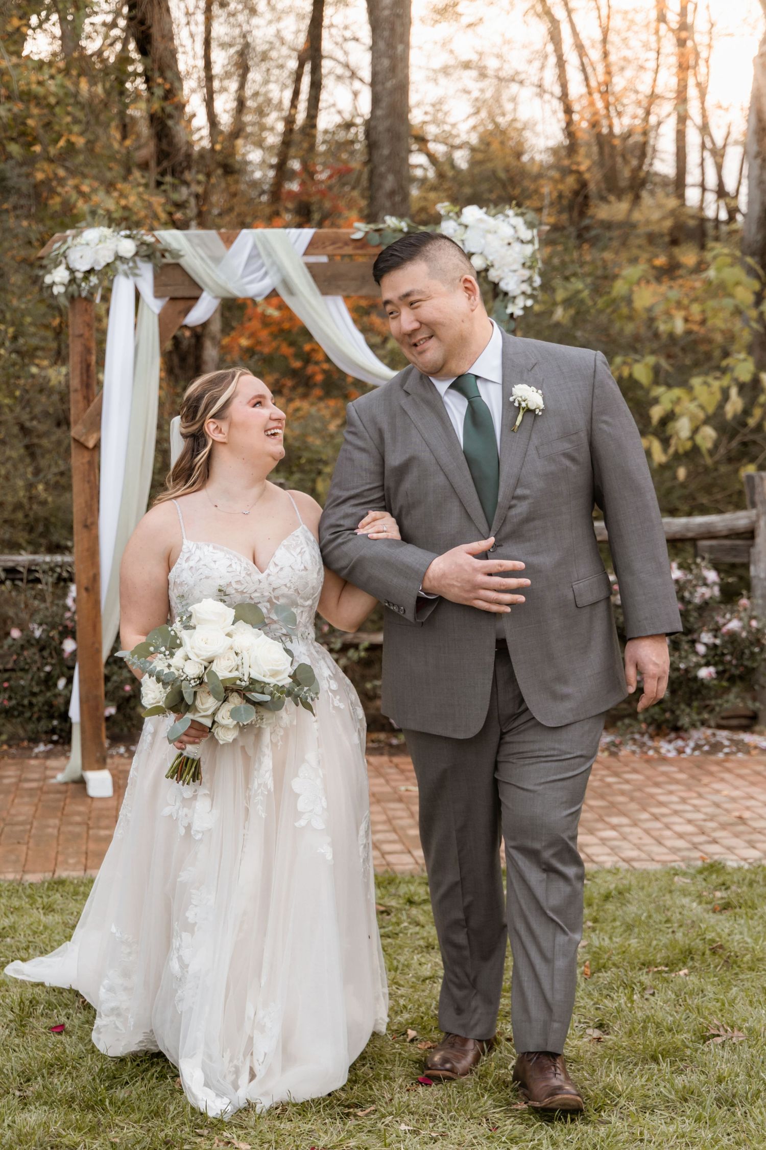 Wedding couple walking together on brick path with white floral arch and autumn foliage in background.