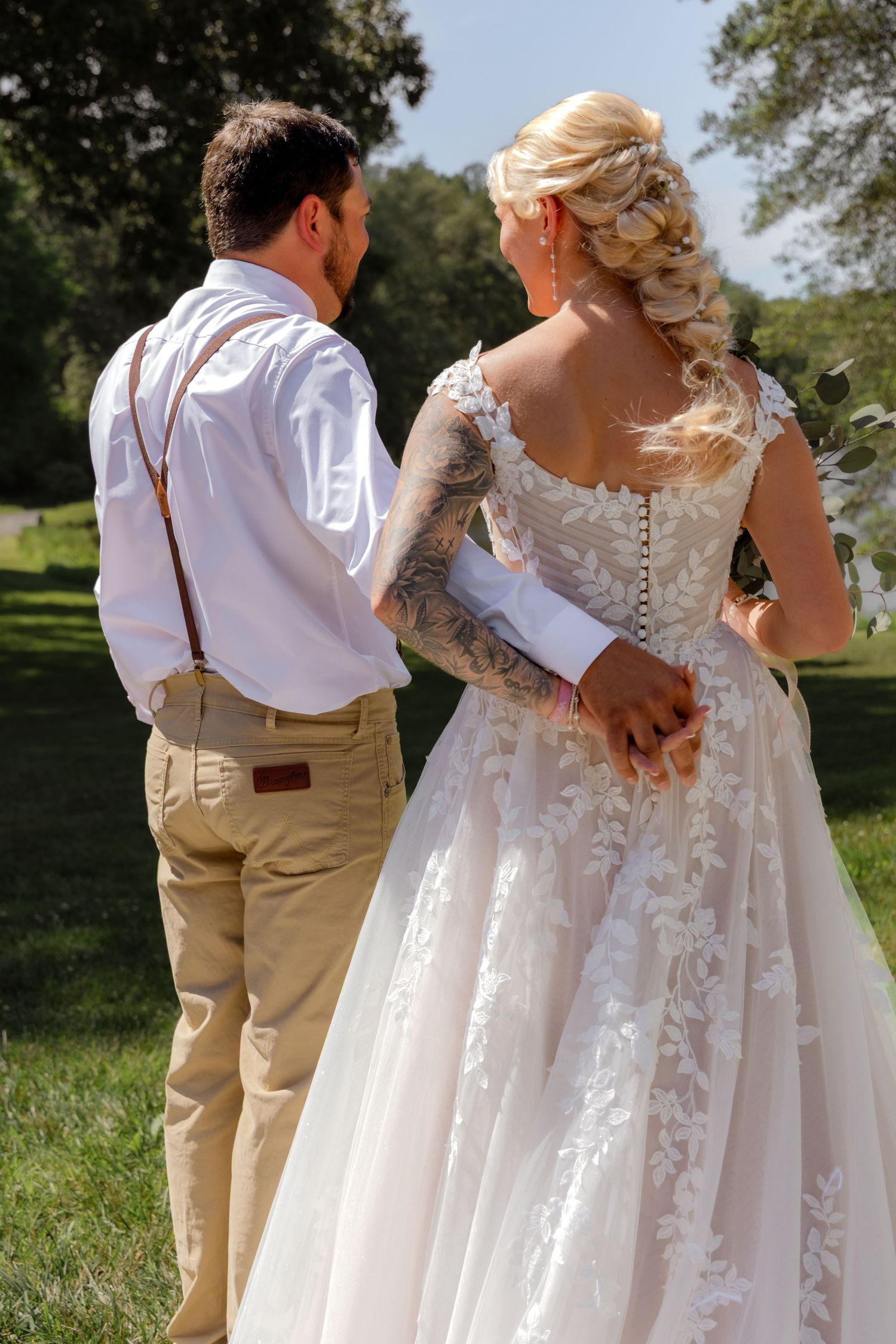 A bride in a lace gown and groom in suspenders walk hand-in-hand through a sunny green park.