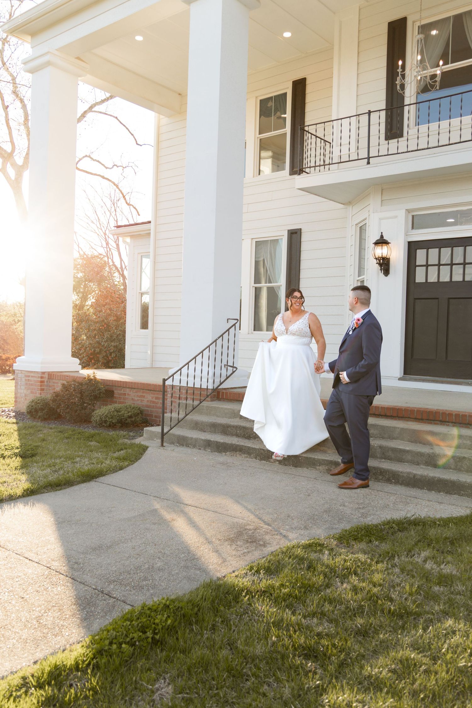 Bride and groom holding hands on steps of elegant white colonial home during golden hour wedding portrait.
