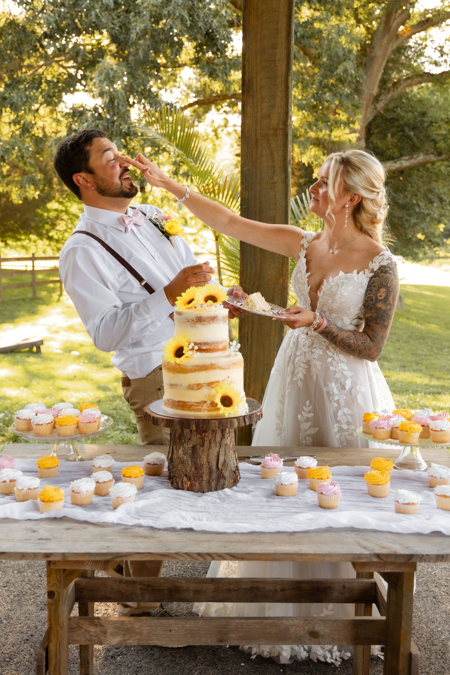 Bride and groom smashing wedding cake into each other faces at rustic outdoor reception with cupcakes.
