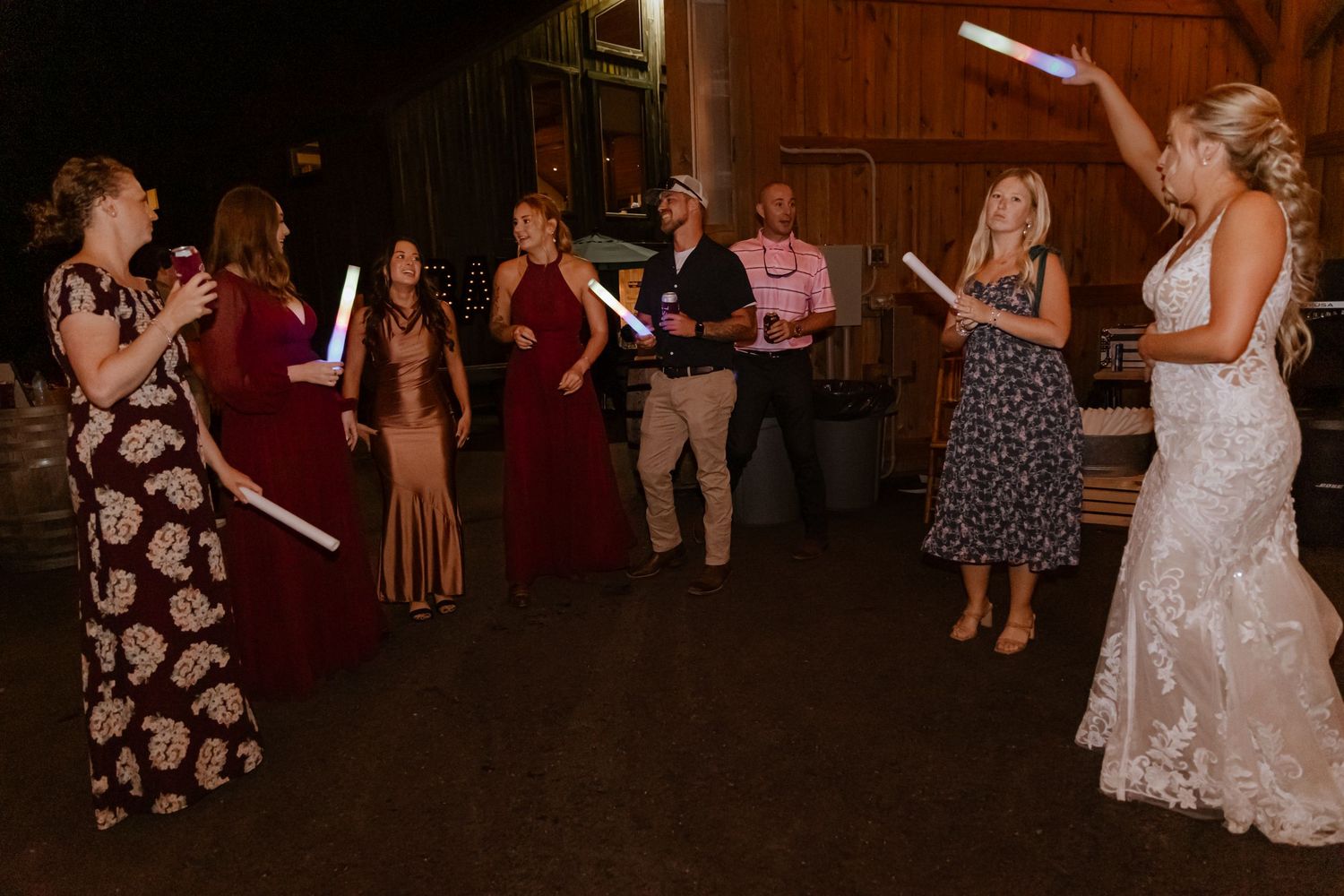 Bride in white gown celebrates with wedding guests holding glowing light sticks at indoor barn reception.