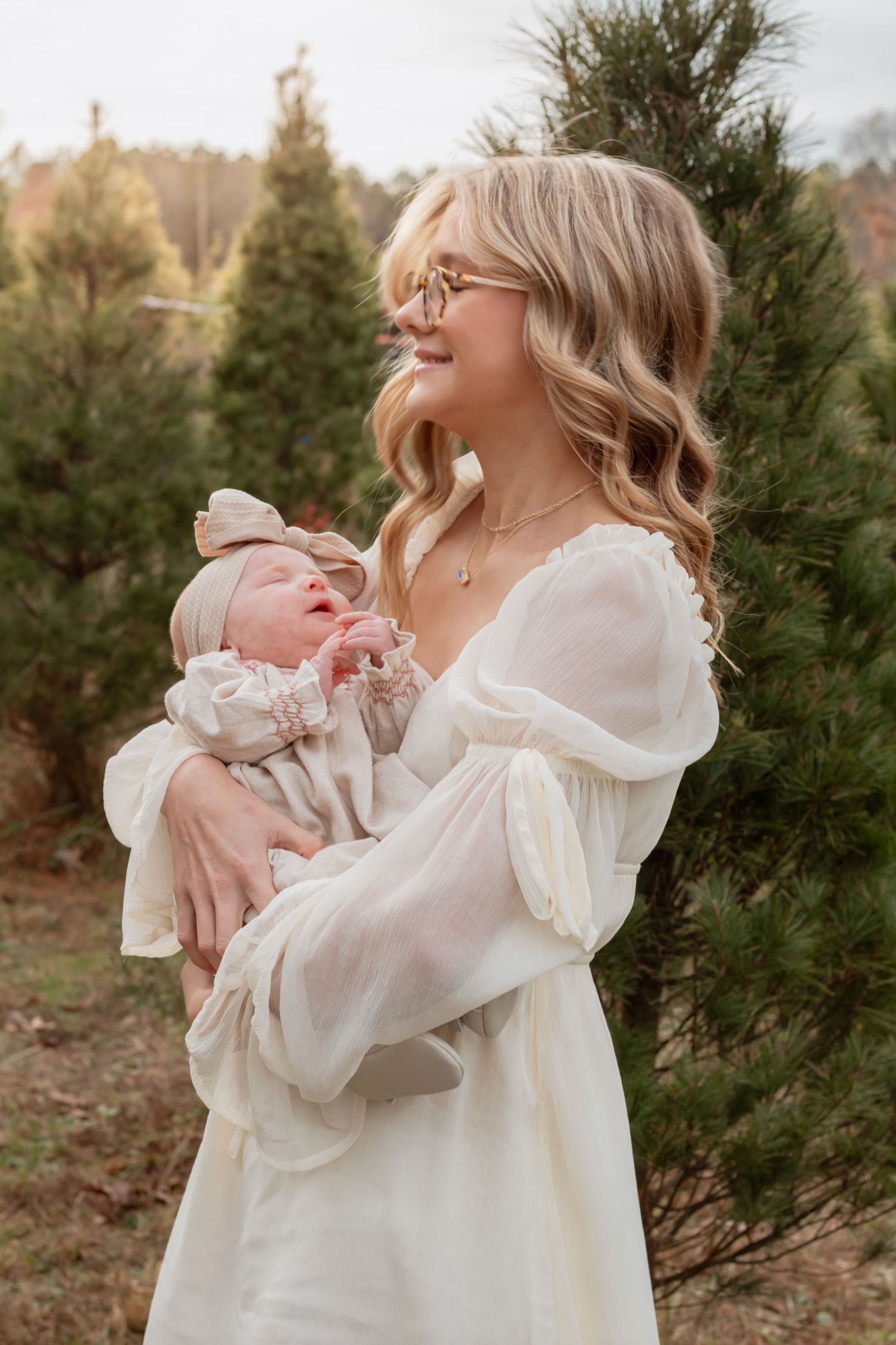 Parent in white dress holding infant outdoors with evergreen trees in background.