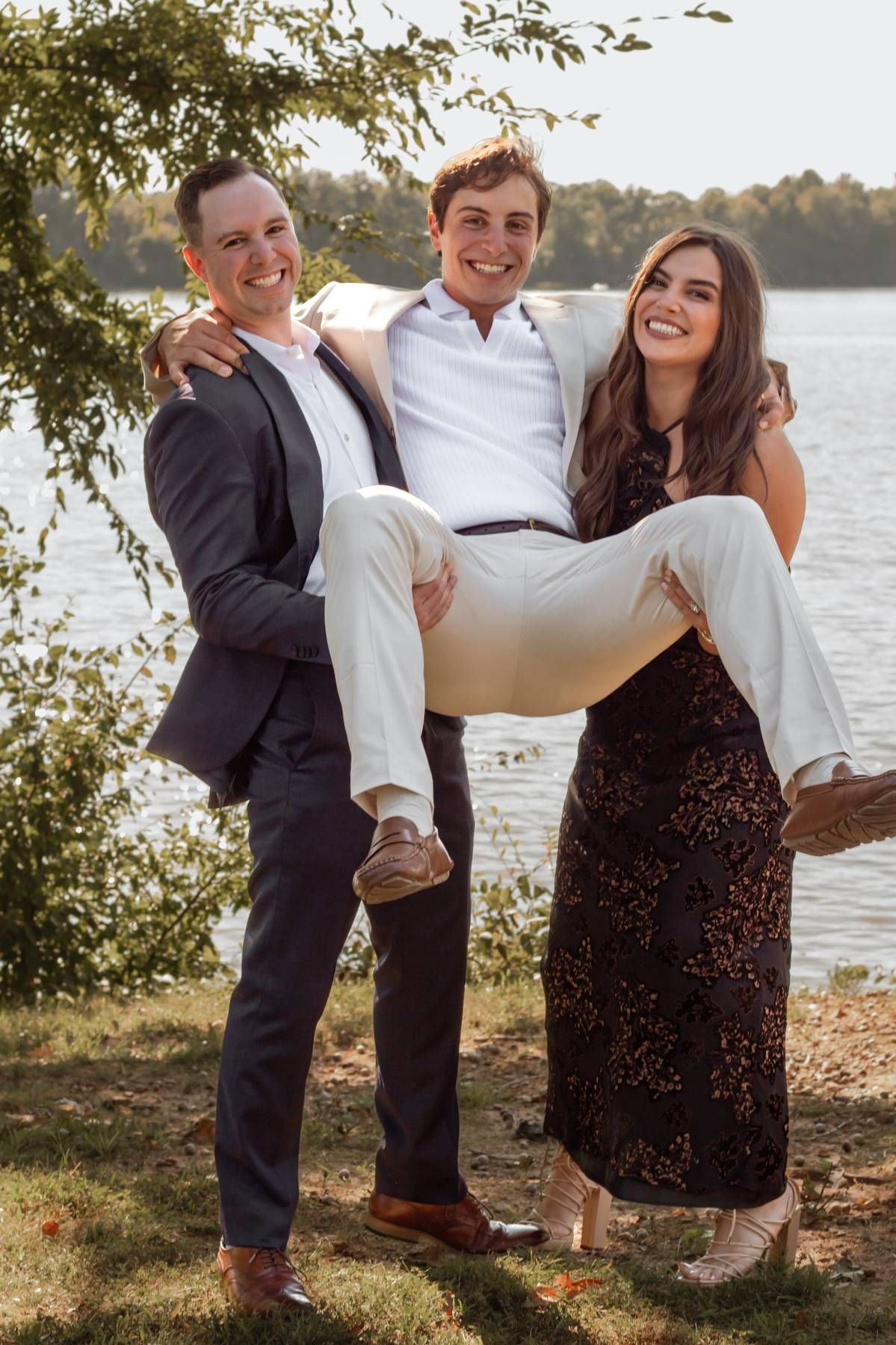 Wedding party members playfully lift someone in formal attire by lakeside.