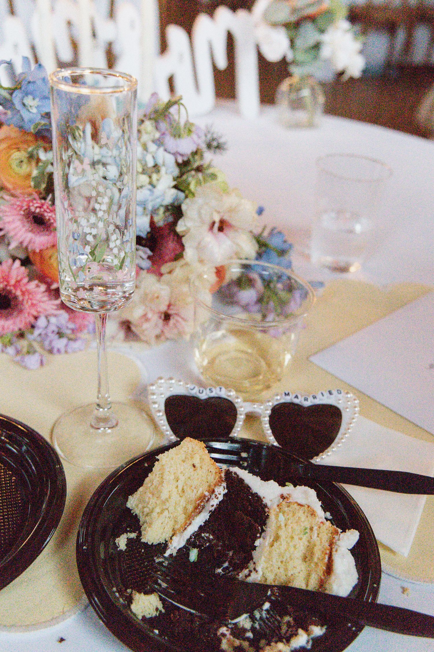 Wedding reception table with layered cake slice, champagne flute, and colorful floral centerpiece on white tablecloth.