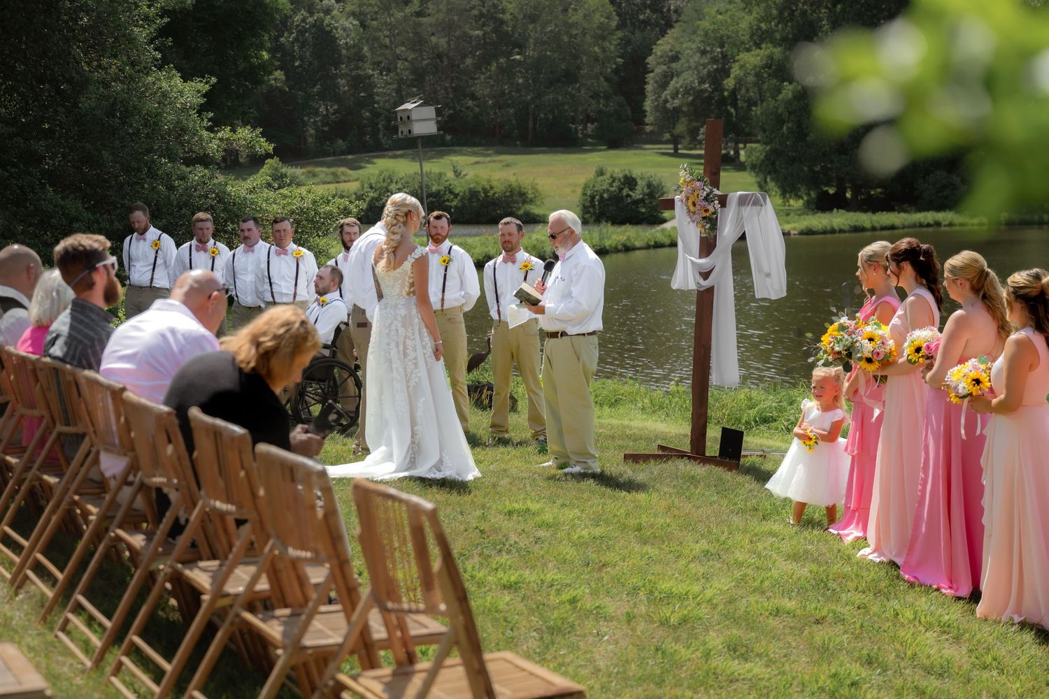 Outdoor wedding ceremony by a pond with bride, groom, bridesmaids in pink, and groomsmen in khaki amid lush greenery.