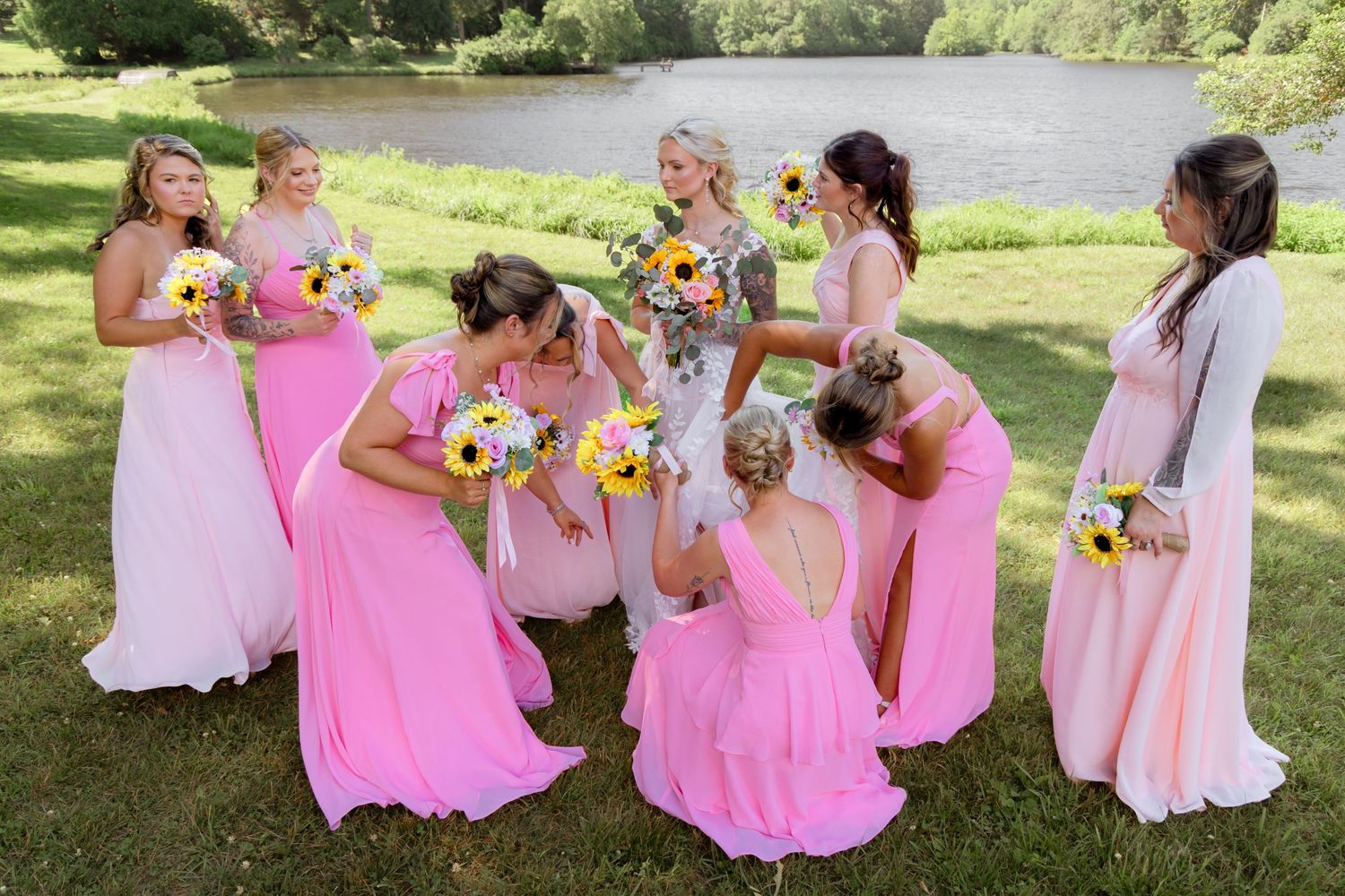 Bridesmaids in pink dresses holding sunflower bouquets gather outdoors near a pond on a sunny wedding day.