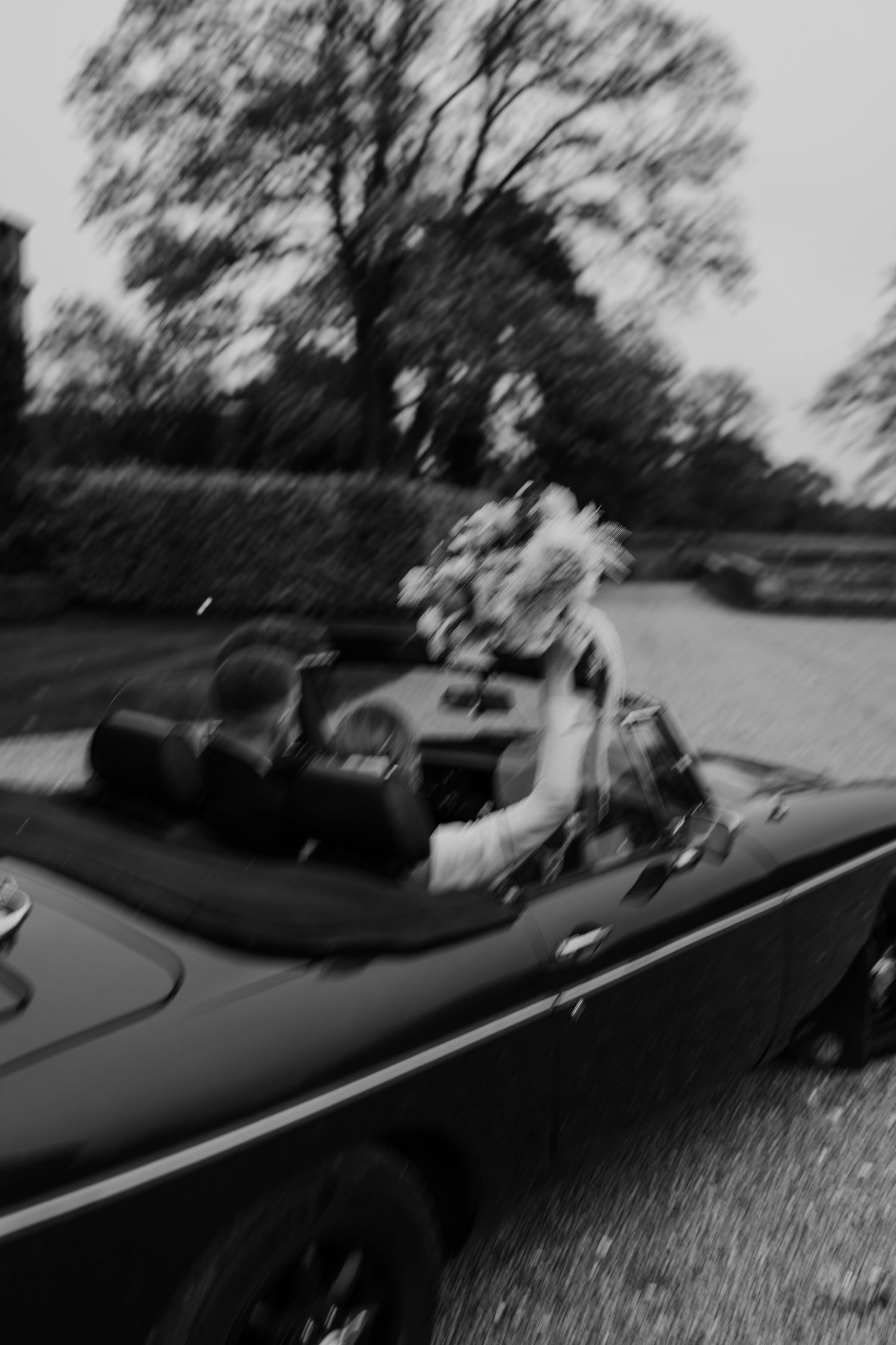 A black and white photograph shows an antique convertible car decorated with flowers driving along a tree-lined road.