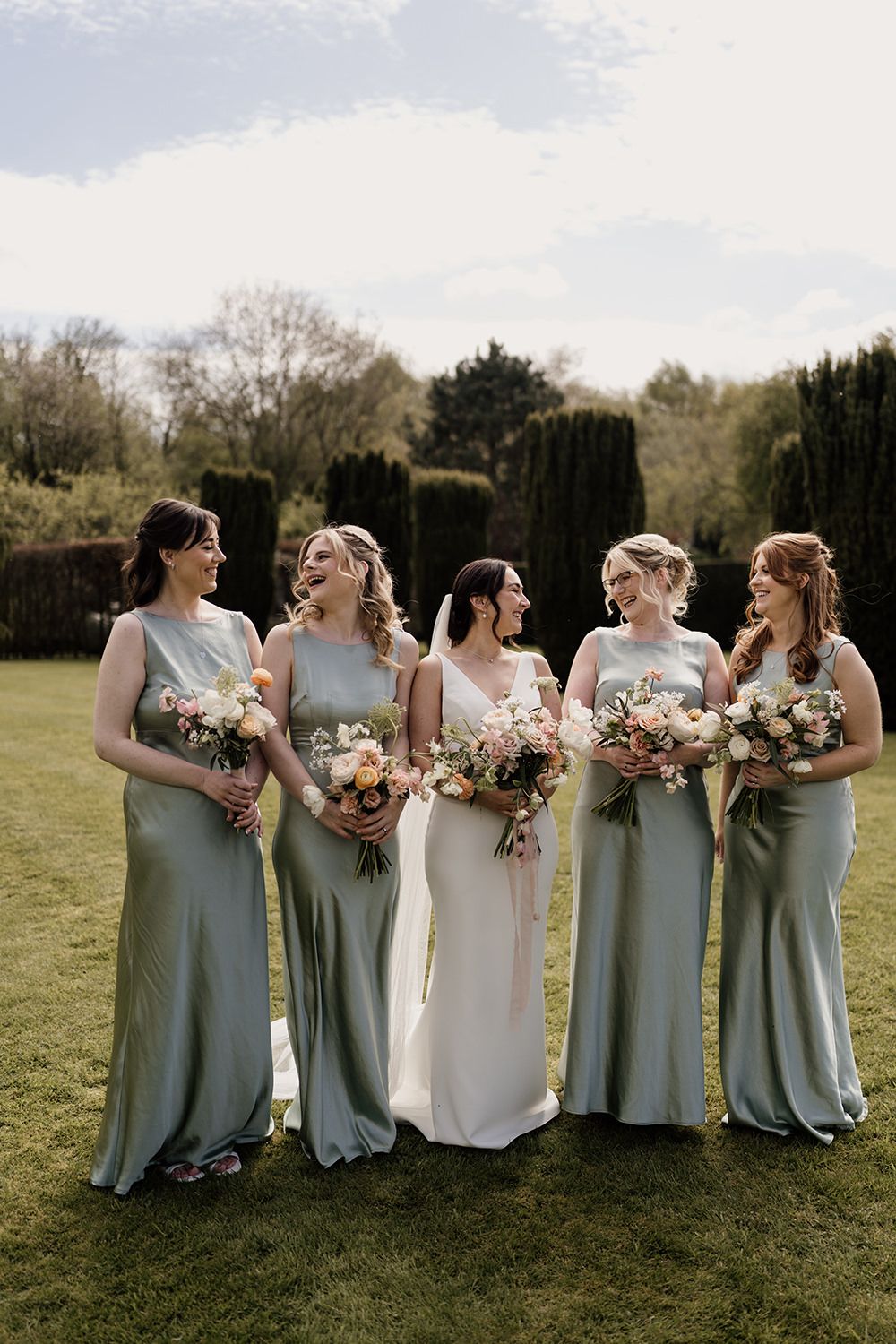 Bride and bridesmaids looking and smiling at each other in the gardens at The Holford Estate.