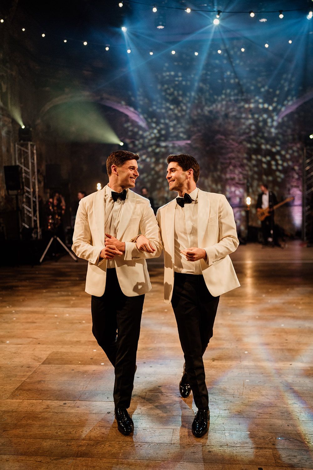 Two grooms in monochrome suits dancing their first dance with stage lights and a band behind them.