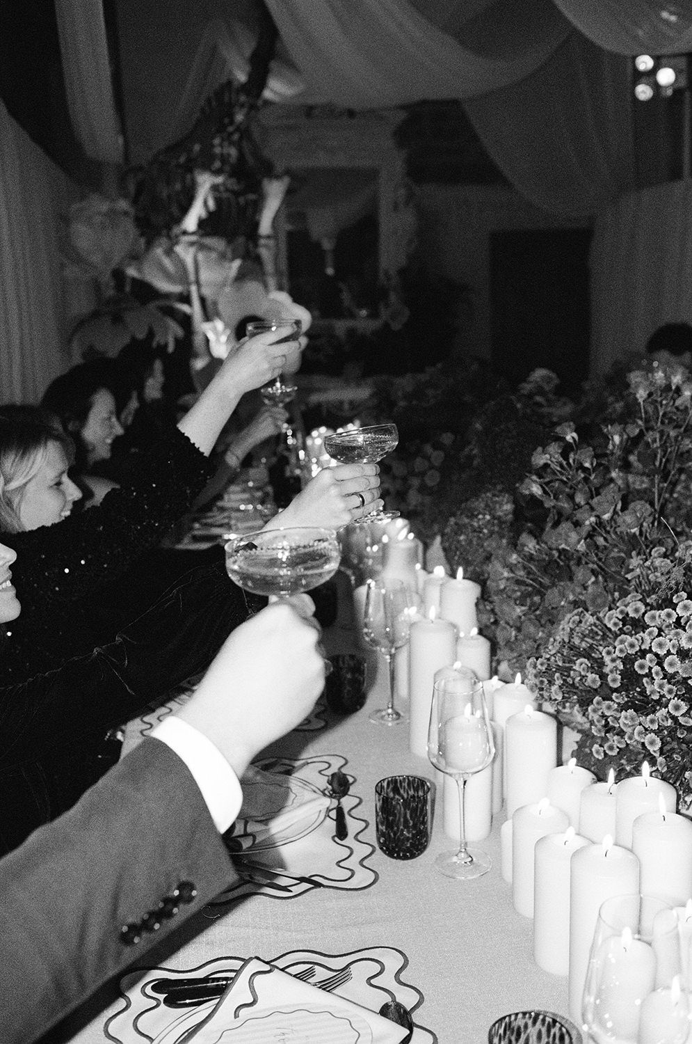 Black and white photo of hands raising champagne glasses at an elegant party with candles and flowers on display.