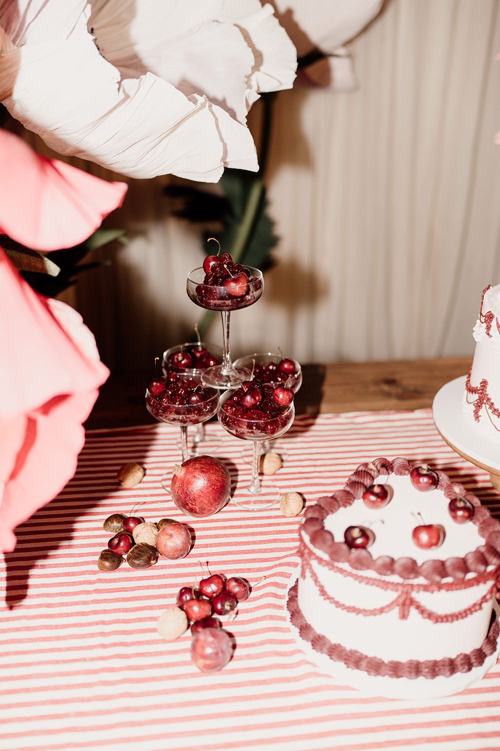 Red and pink holiday table setting with tiered dessert stand, cherries, ornaments and cake on striped tablecloth.