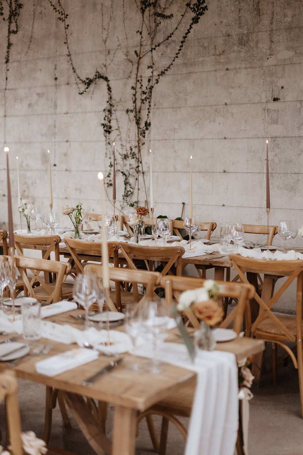 Rustic wedding reception table setting with wooden cross-back chairs, white linens, and delicate floral arrangements.