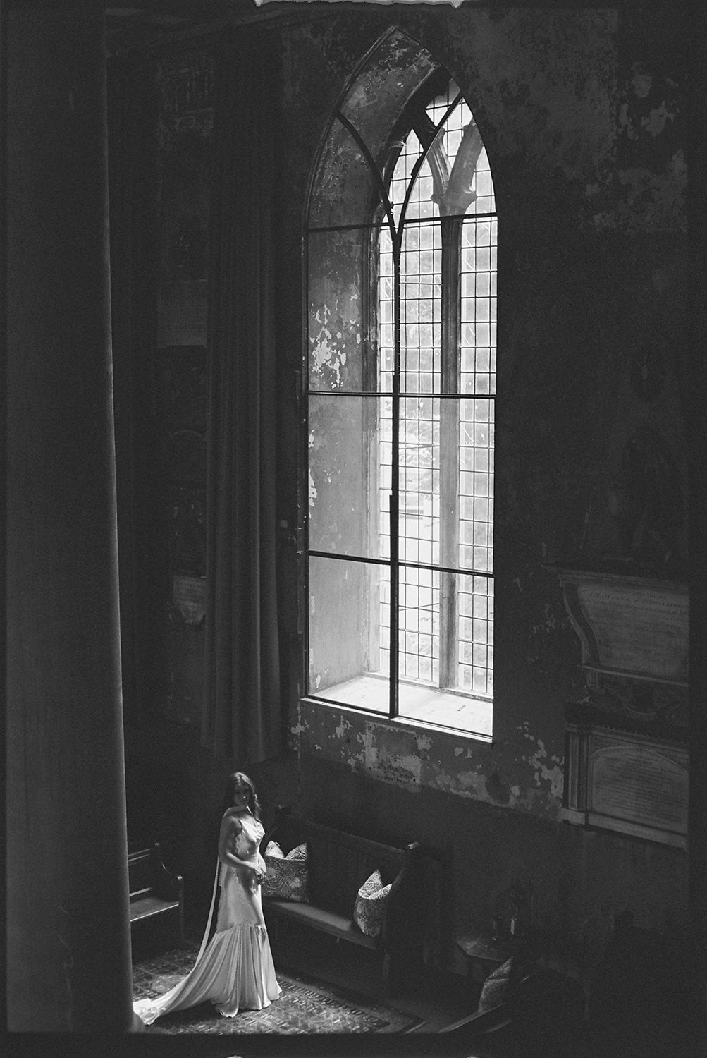 A figure in a flowing dress stands near Gothic windows in a dark, atmospheric church interior.