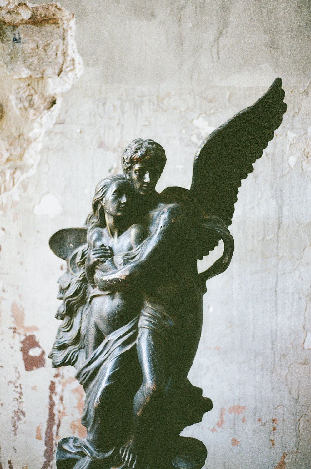 A dramatic stone sculpture of an angel embracing a figure against a weathered white wall creates a moody artistic scene.