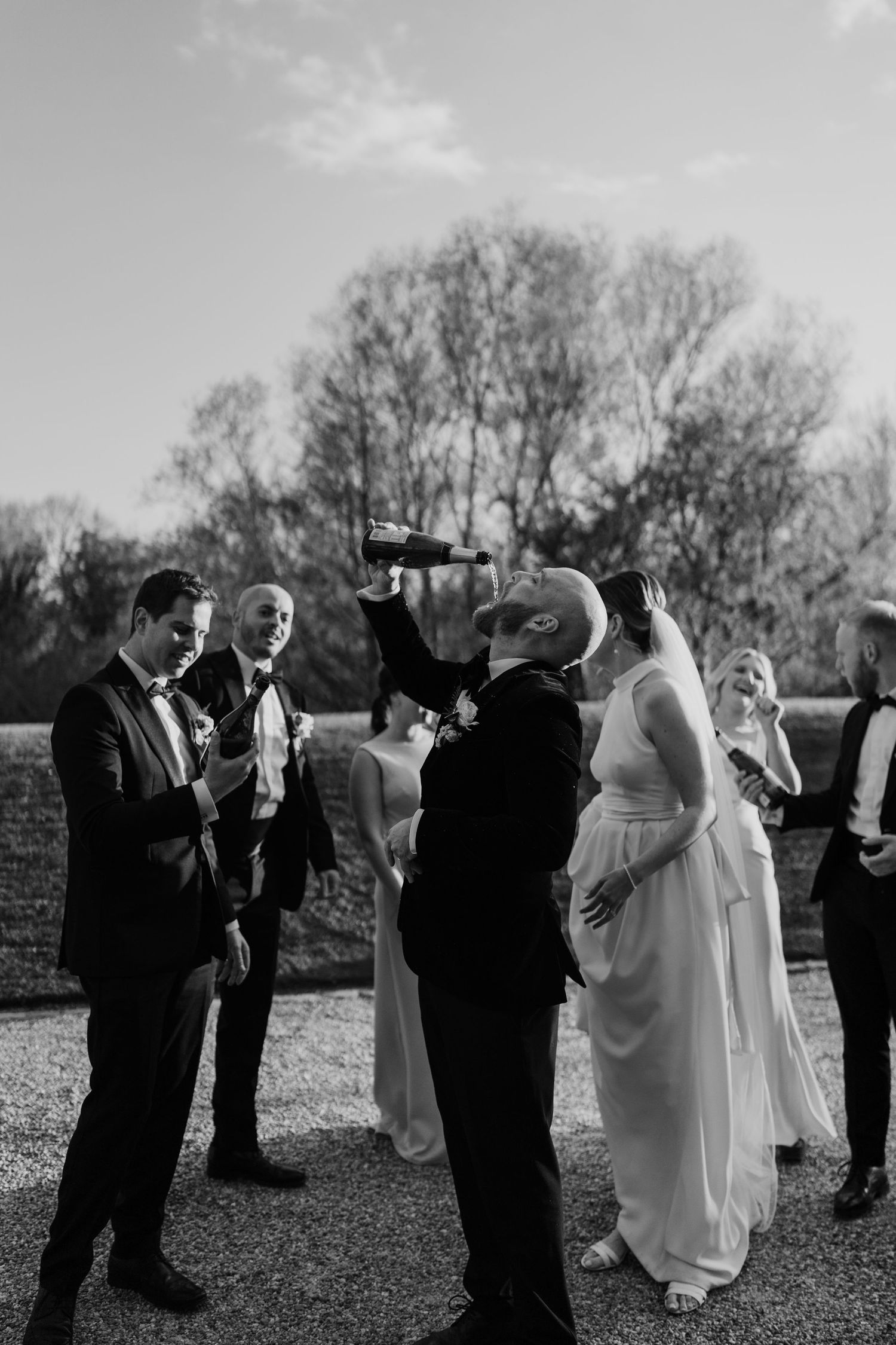 Wedding guests gather outdoors for a ceremony on a sunny day with bare trees in the background.