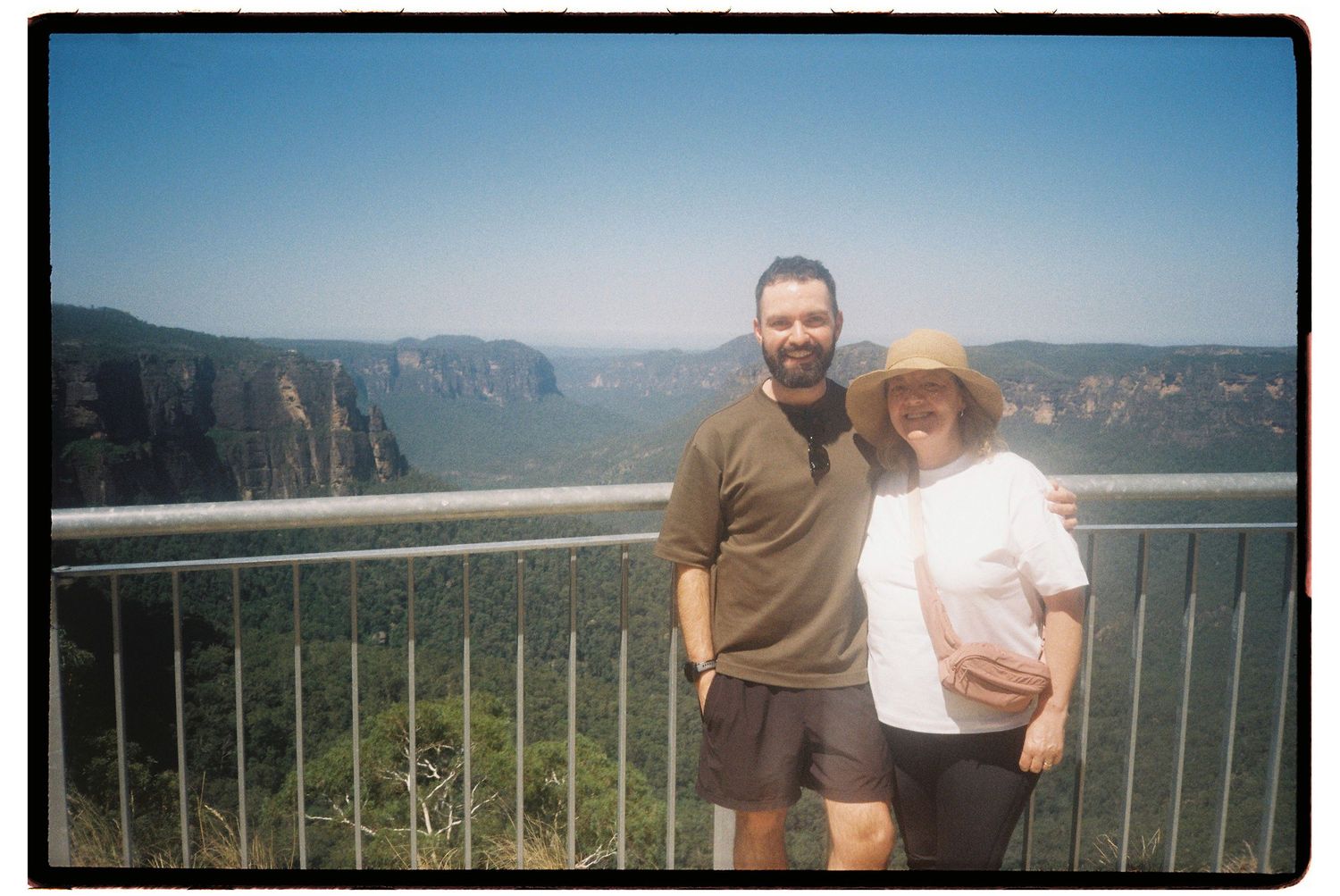 Couple smiling at scenic mountain valley overlook on sunny day.