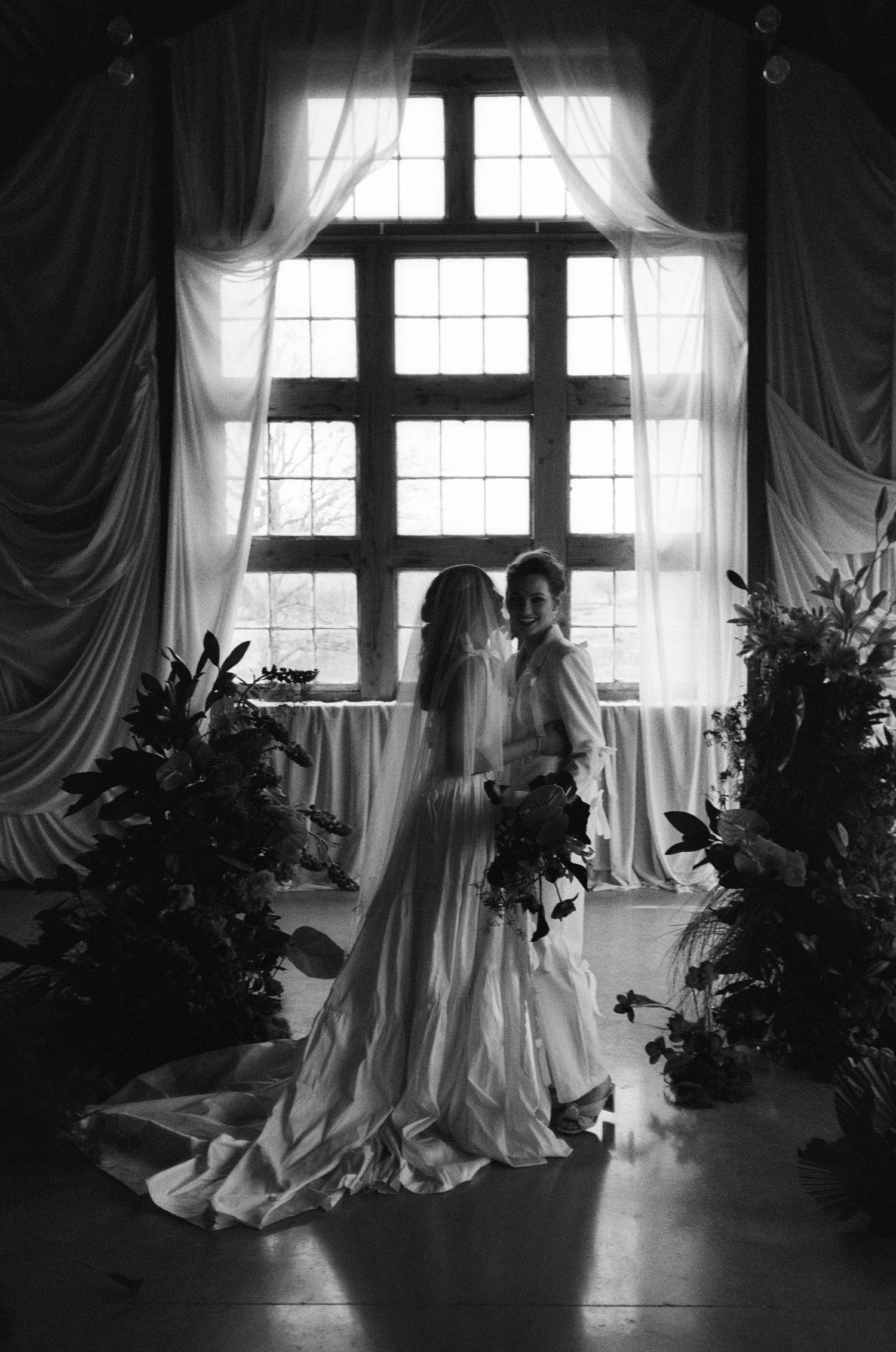Black and white wedding photo of two brides standing before a large window surrounded by lush greenery.
