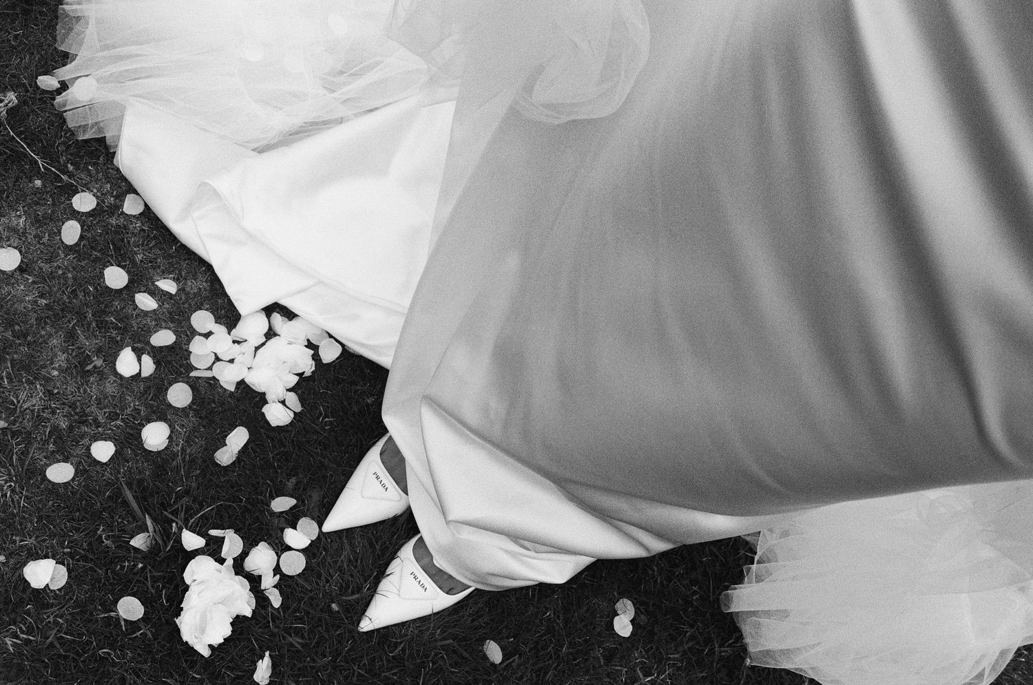 Black and white photo of a bride's white gown and heels surrounded by scattered flower petals on grass.