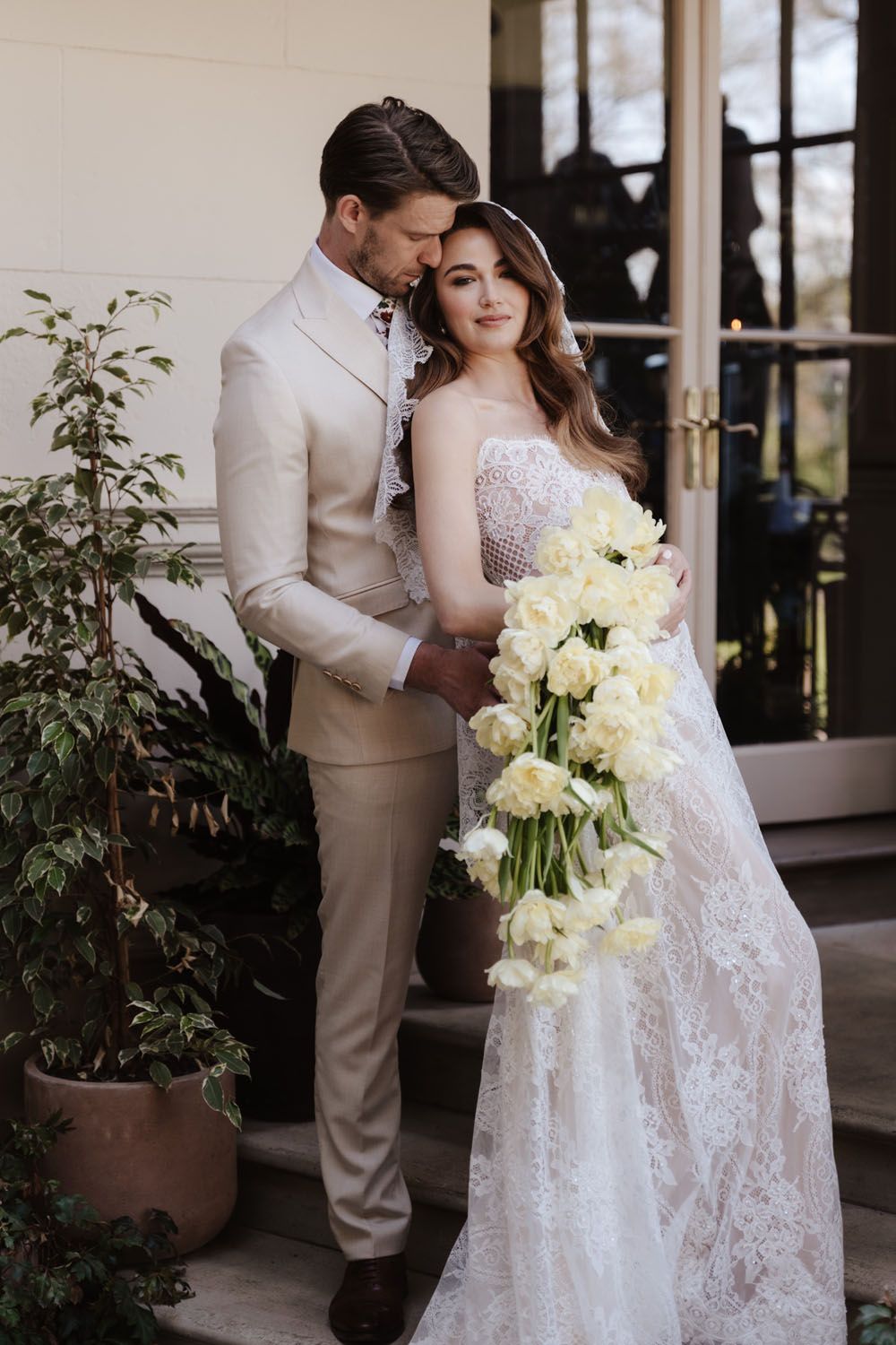 Bride in lace gown holding white tulip bouquet embraced by groom in beige suit outside elegant venue.