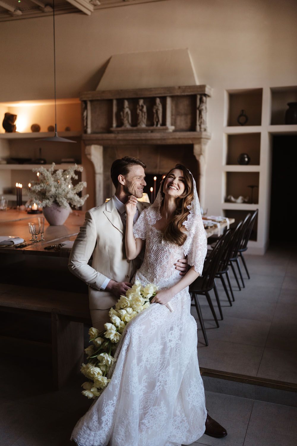 Bride in lace gown holding white bouquet stands with groom in beige suit near rustic fireplace in elegant wedding venue.