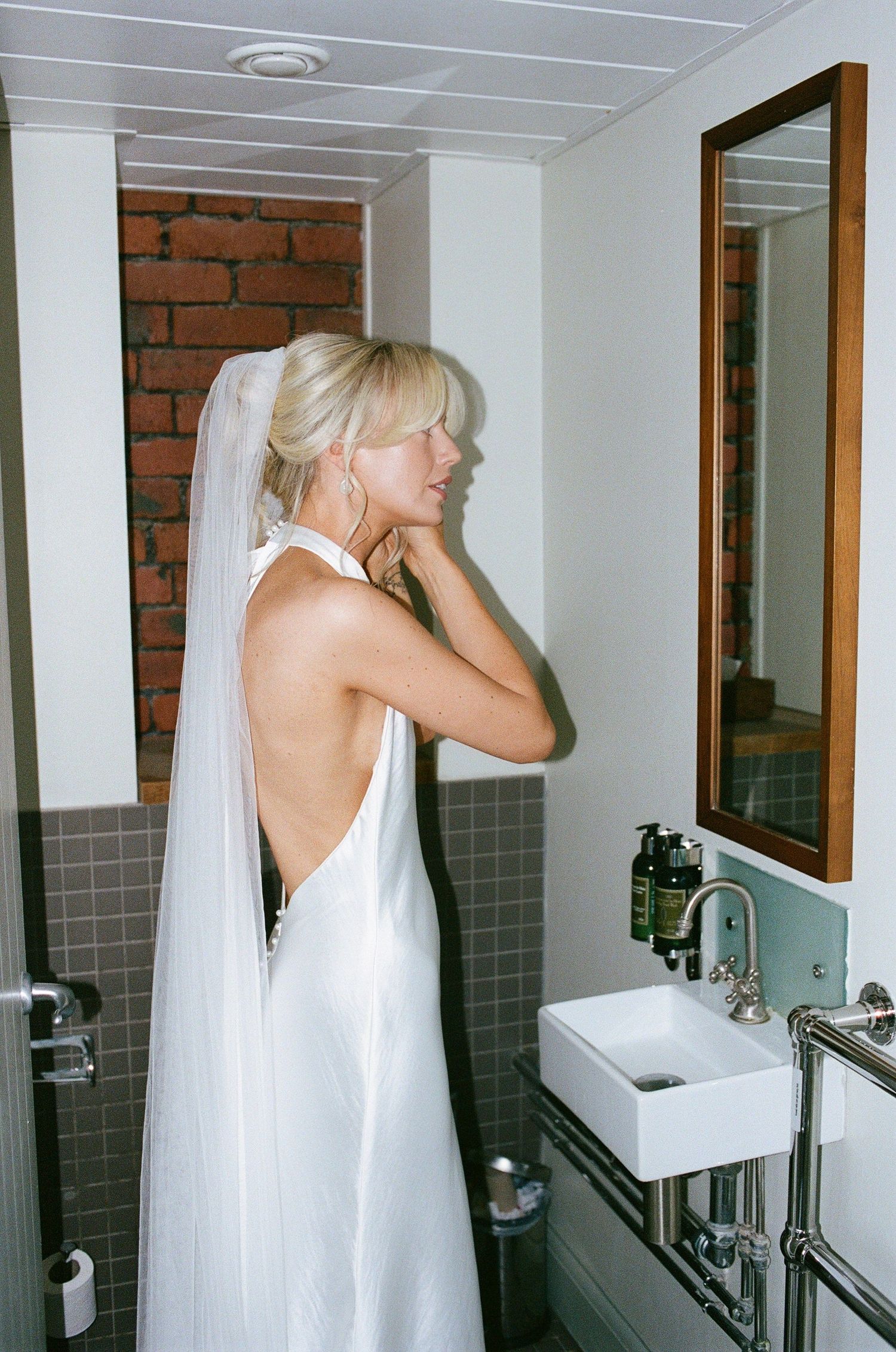 A person in a white halter dress stands in a modern bathroom with exposed brick walls and a framed mirror.
