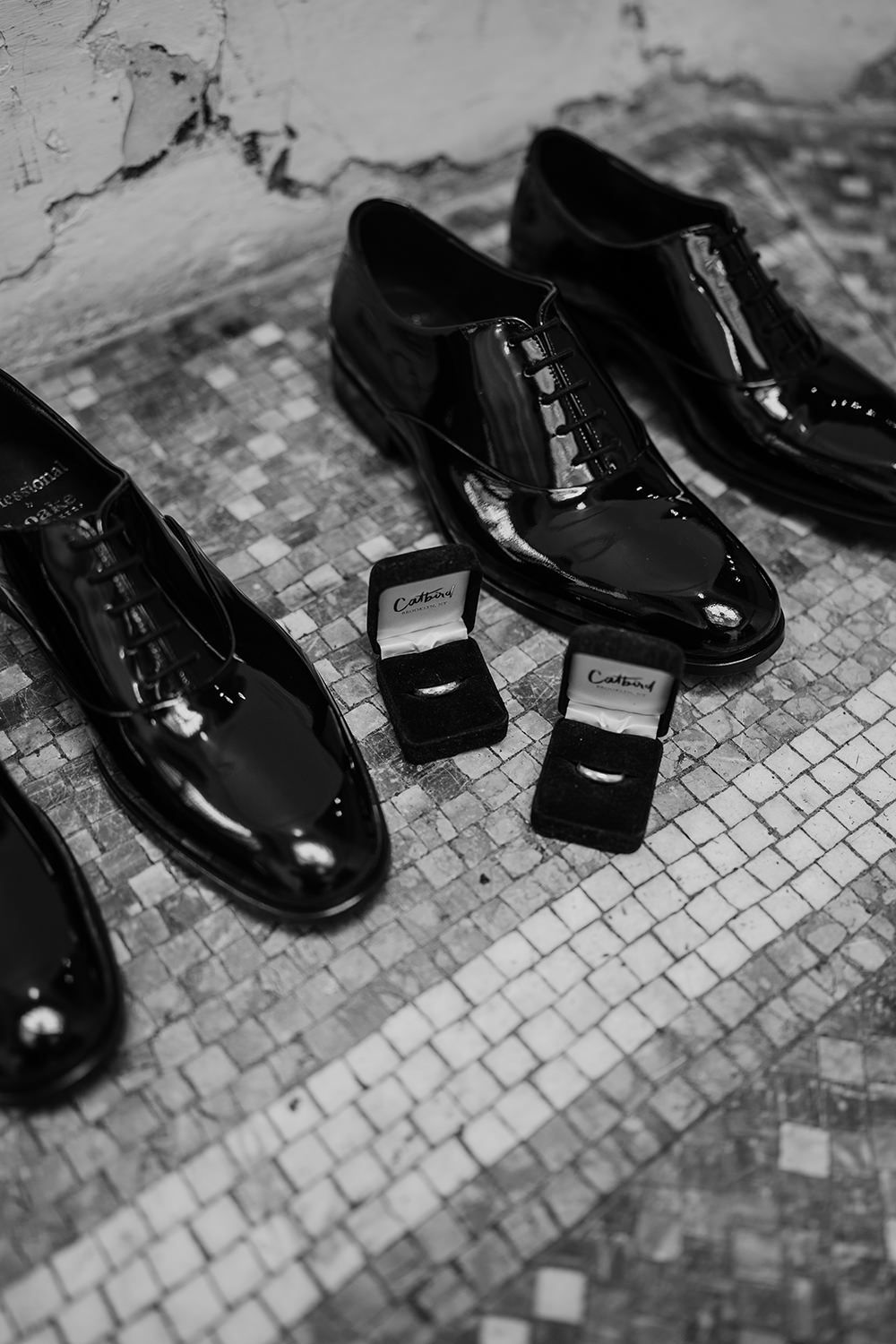 Two pairs of black shiny wedding shoes with two wedding ring boxes beteen, both with rings inside.