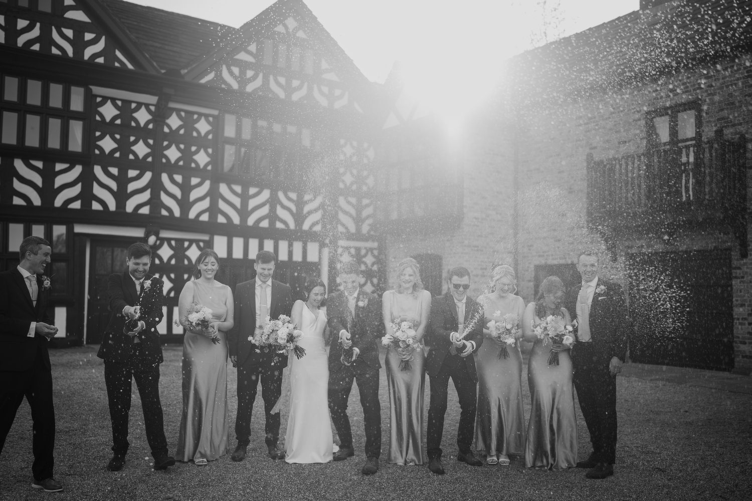 Bride and Groom standing with a bottle of champagne spraying surrounded by their wedding party with Holford Hall behind.
