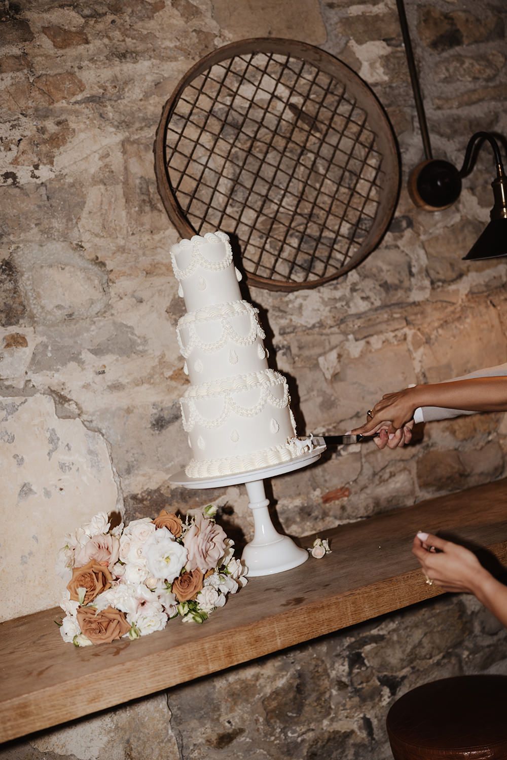 A white tiered wedding cake sits on a wooden surface with romantic floral decorations beneath an industrial metal grate.