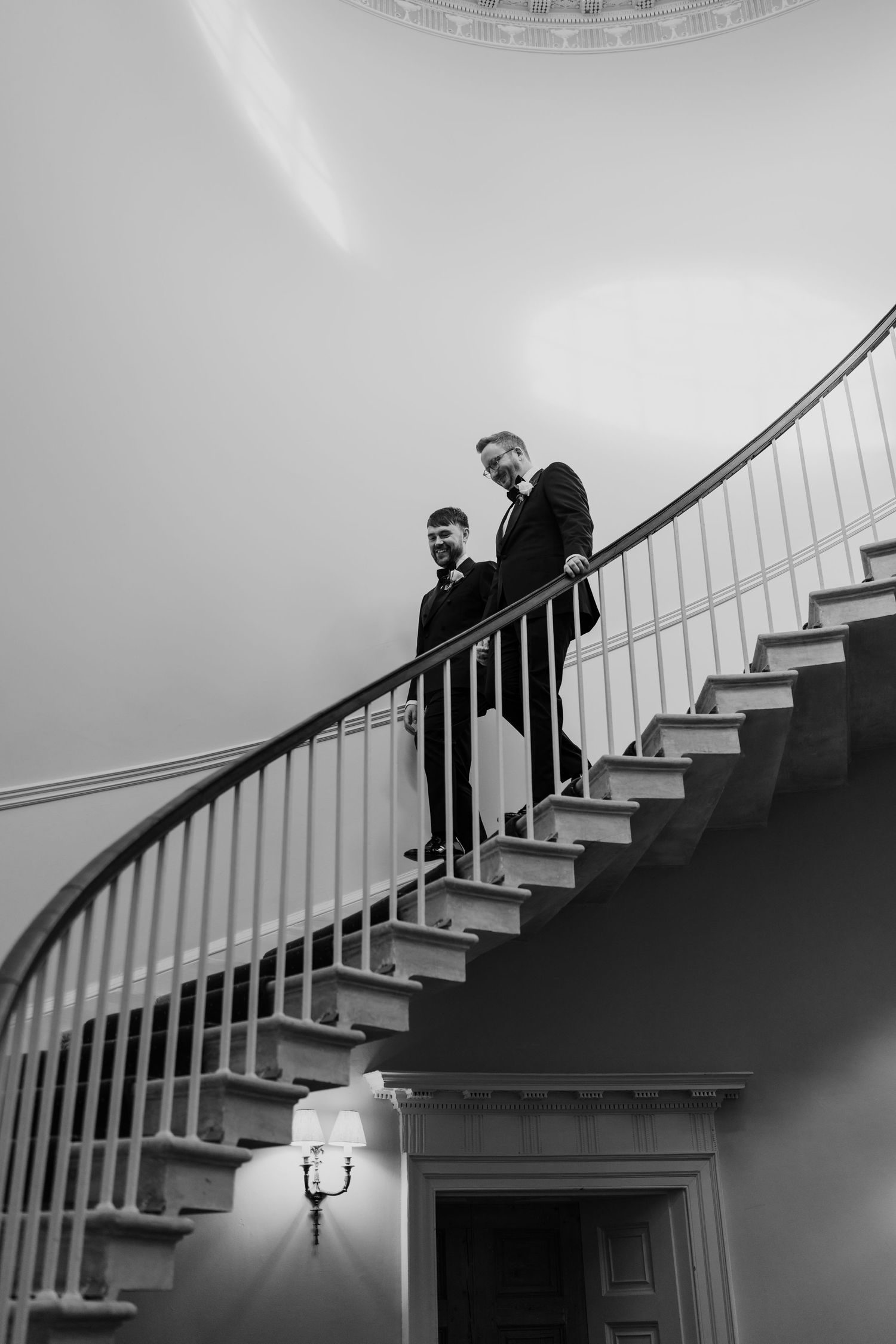 Two grooms in formal attire stand on an elegant curved staircase in a black and white wedding photo.