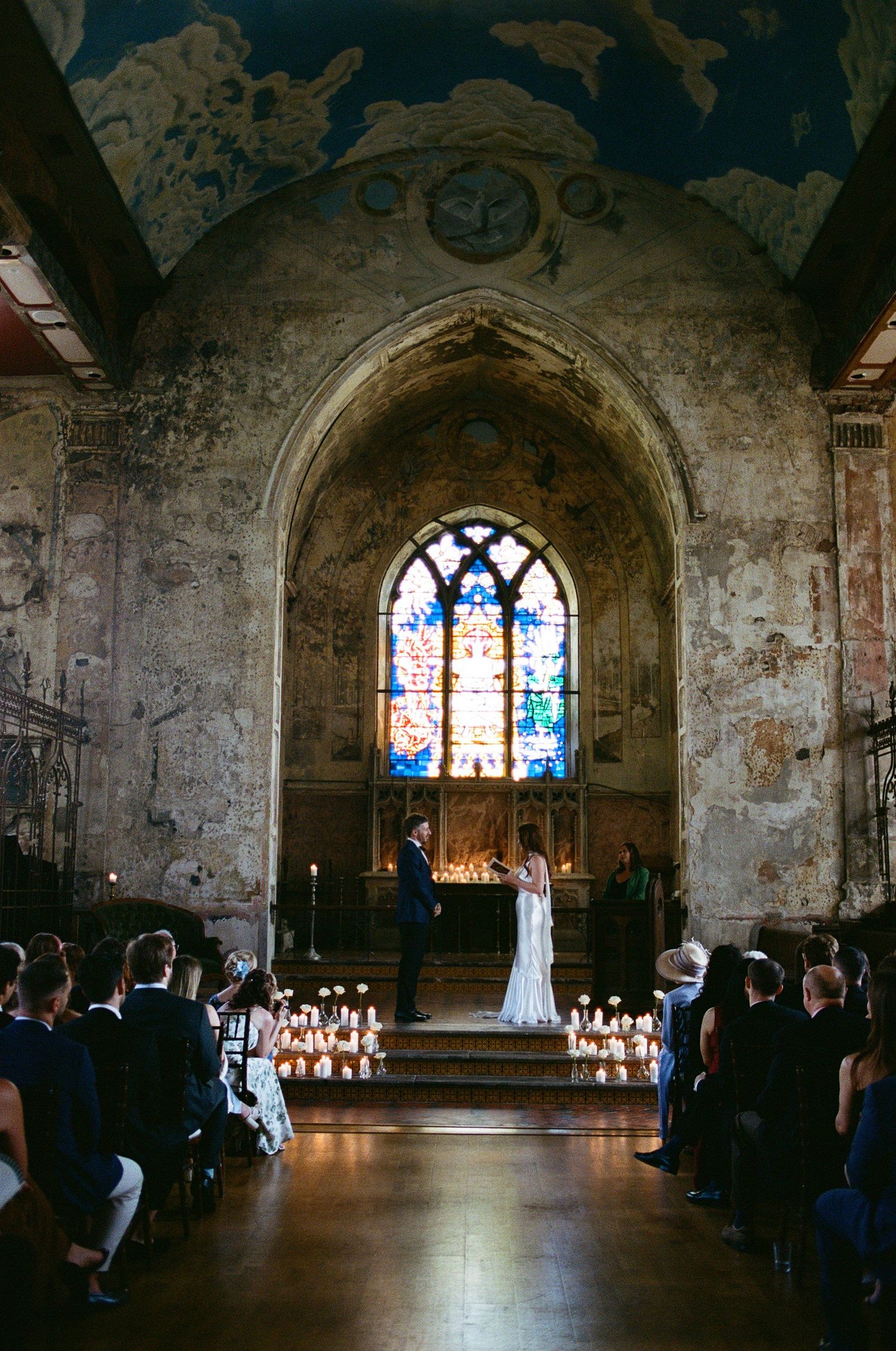 Romantic wedding ceremony in a historic church with stained glass windows, candlelit altar, and guests seated in wooden pews.