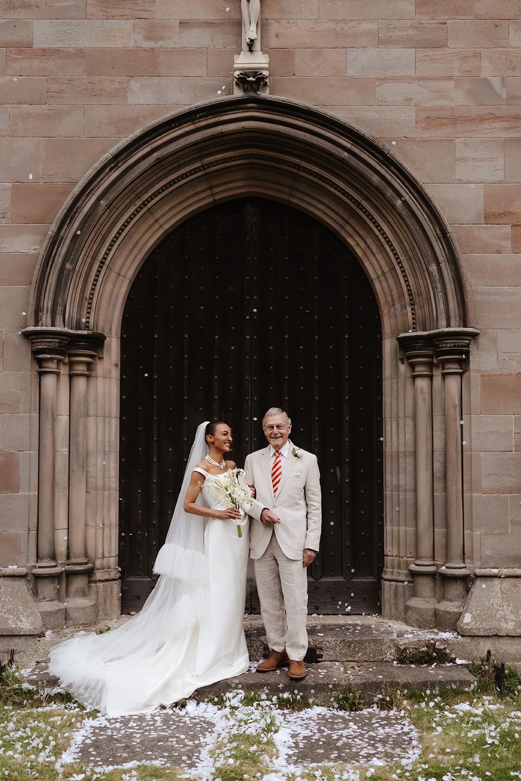 Bride in white gown and veil stands with father at ornate church doorway with confetti falling.