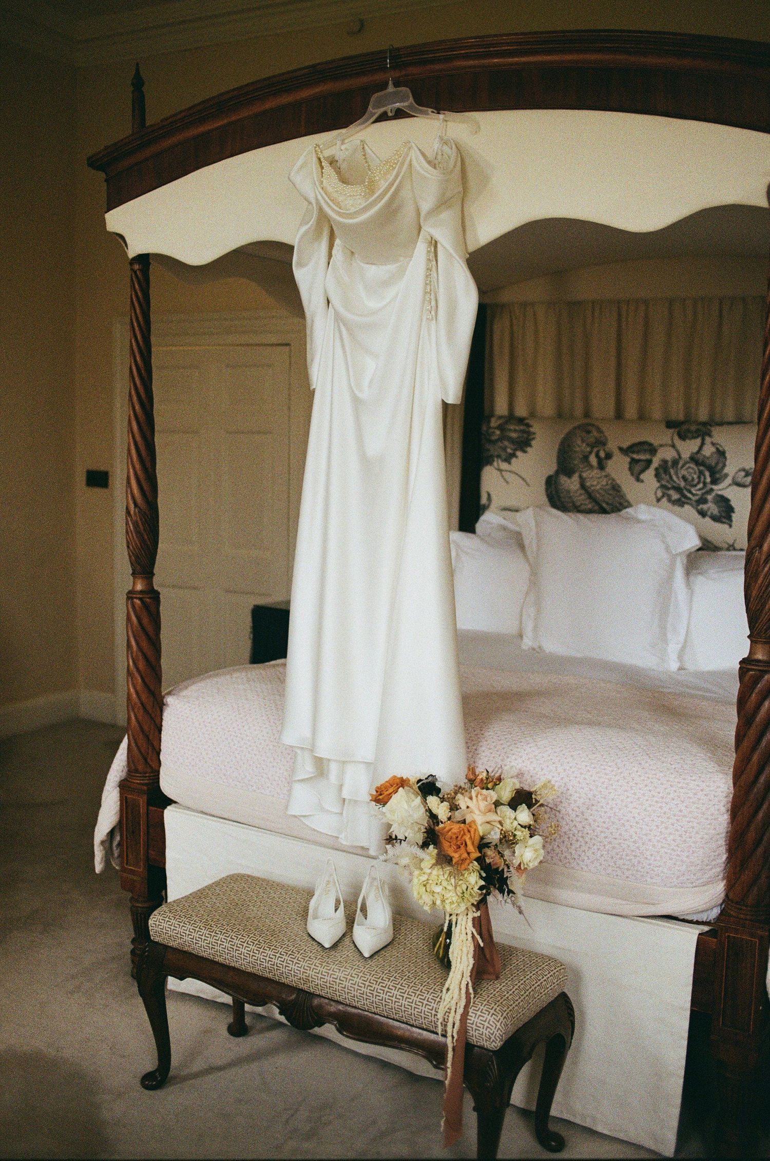 Elegant white wedding dress hangs on four-poster bed with floral bouquet and white shoes displayed on bench below.
