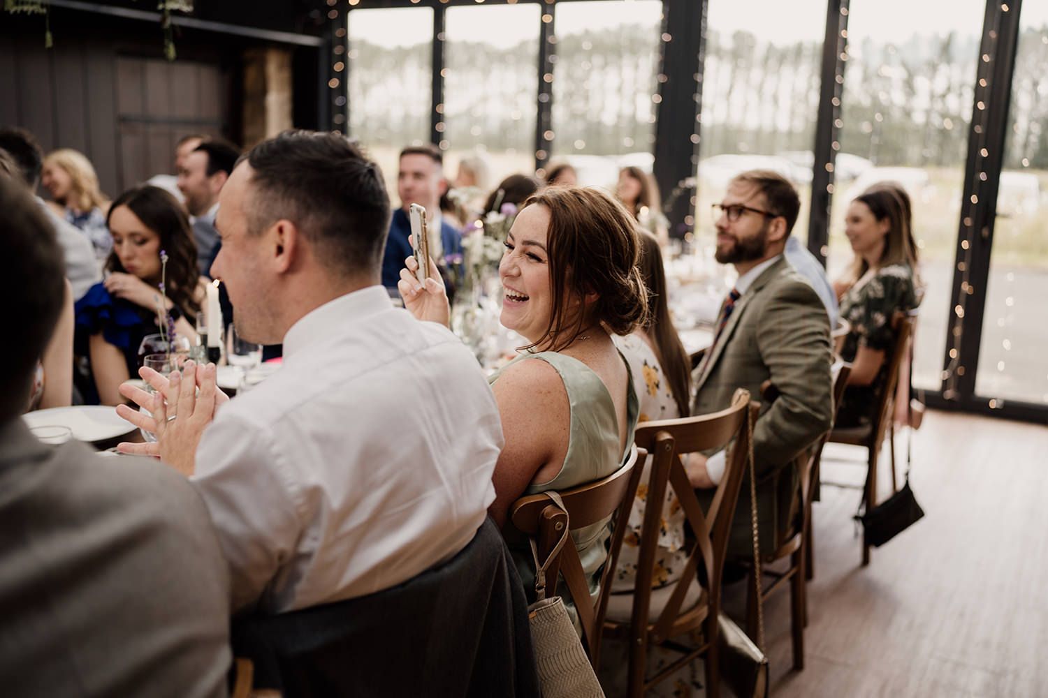 Bridesmaid in green laughing and clapping while sat down during wedding speeches.