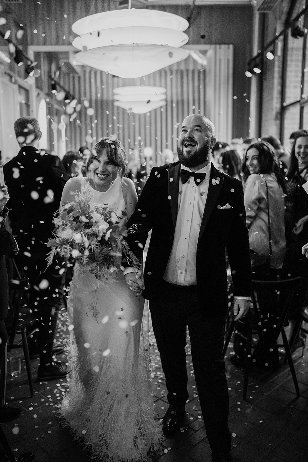 Black and white wedding photo showing joyful couple walking through confetti under modern pendant lights during ceremony.
