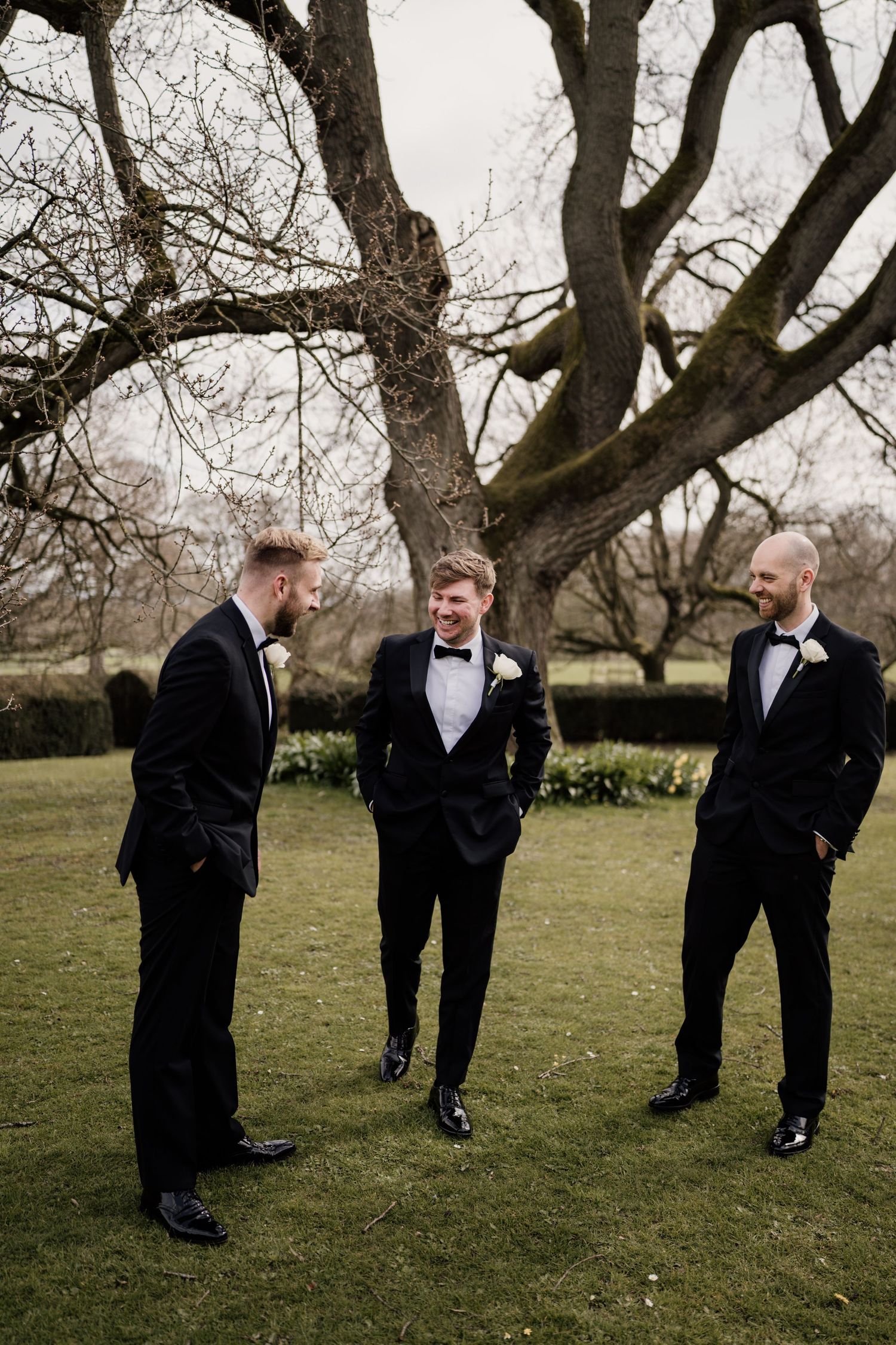 Groomsmen talking outside on grass with trees in the background.
