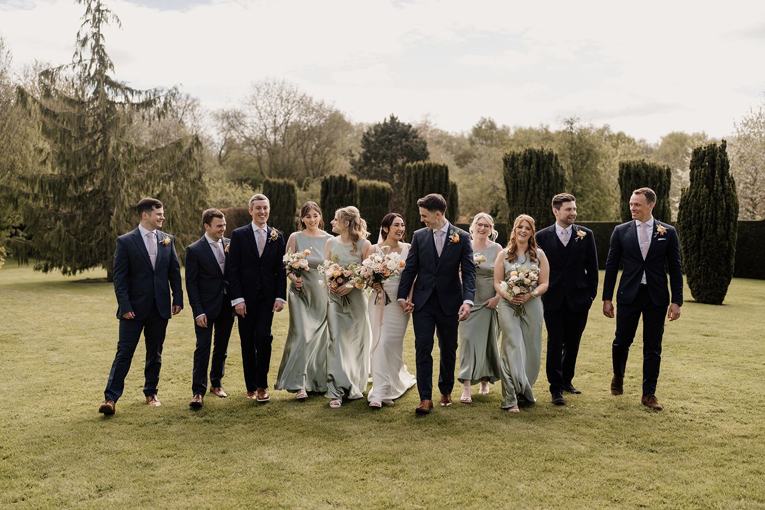 Groomsmen and Bridesmaids walking in a line laughing and talking through the gardens at The Holford Estate.
