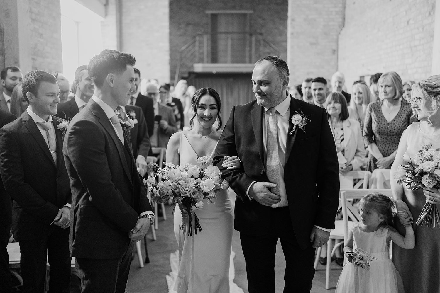 Bride linking arms with her father as she walks down the aisle on her wedding with guests watching at either side.