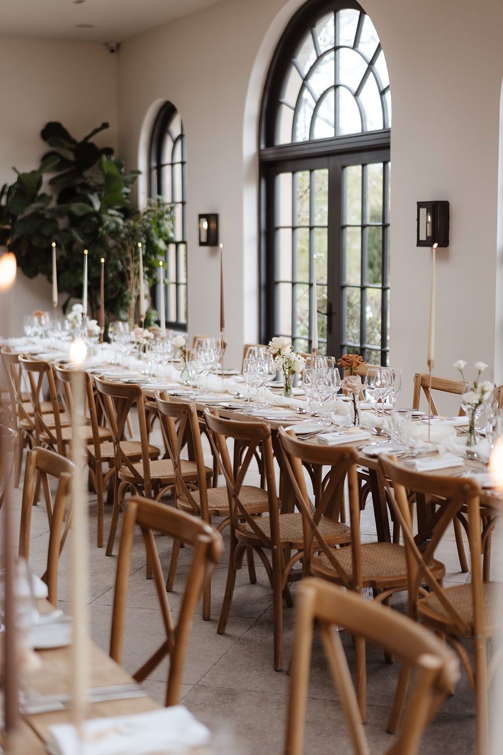 Elegant wedding reception table setting with wooden crossback chairs and arched windows in a bright white venue.
